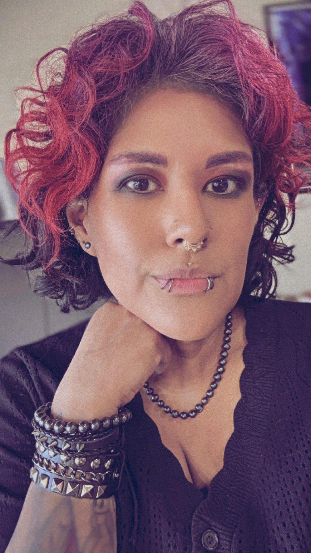 Woman with red curly hair and piercings, wearing a black top and studded bracelet, looking at the camera.