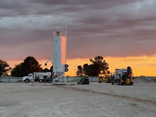 A group of construction vehicles are parked in a gravel lot at sunset.