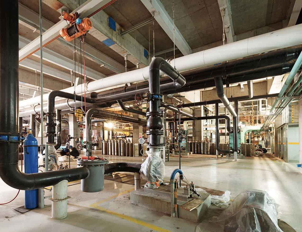 Industrial plant interior with large white pipes, valves, and machinery in a bright factory space
