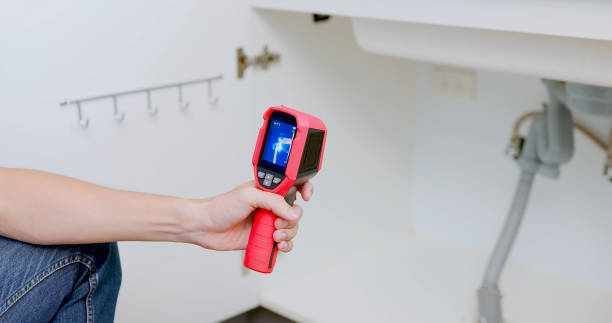 Hand holding a red thermal camera aimed at a white wall in a room
