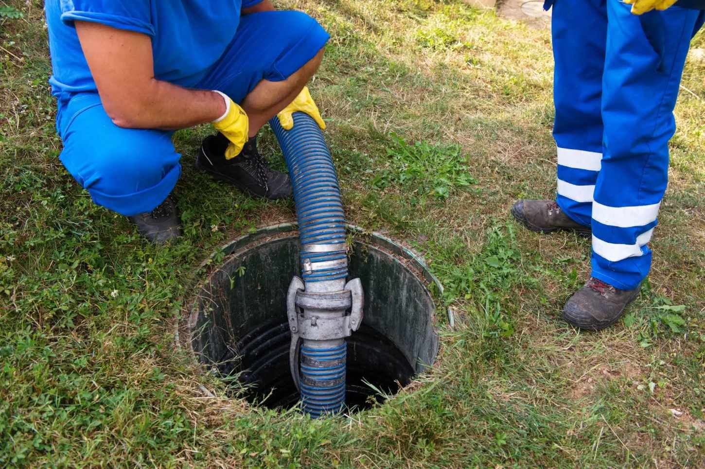 Two workers in blue uniforms handle a hose over an open manhole in grass.
