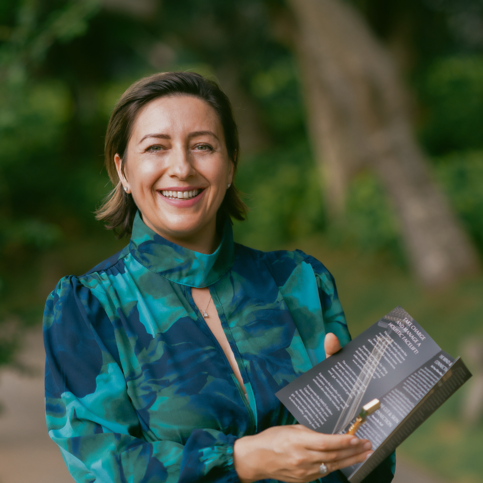Smiling woman outdoors holding a black booklet, wearing a blue-green patterned top.