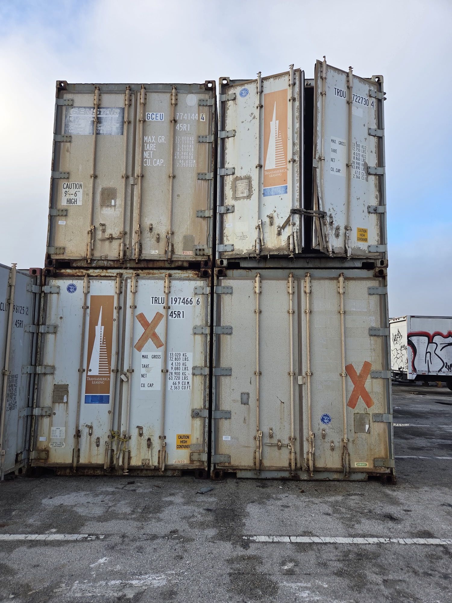 Four weathered shipping containers stacked, one door open, in an outdoor lot against a cloudy sky.