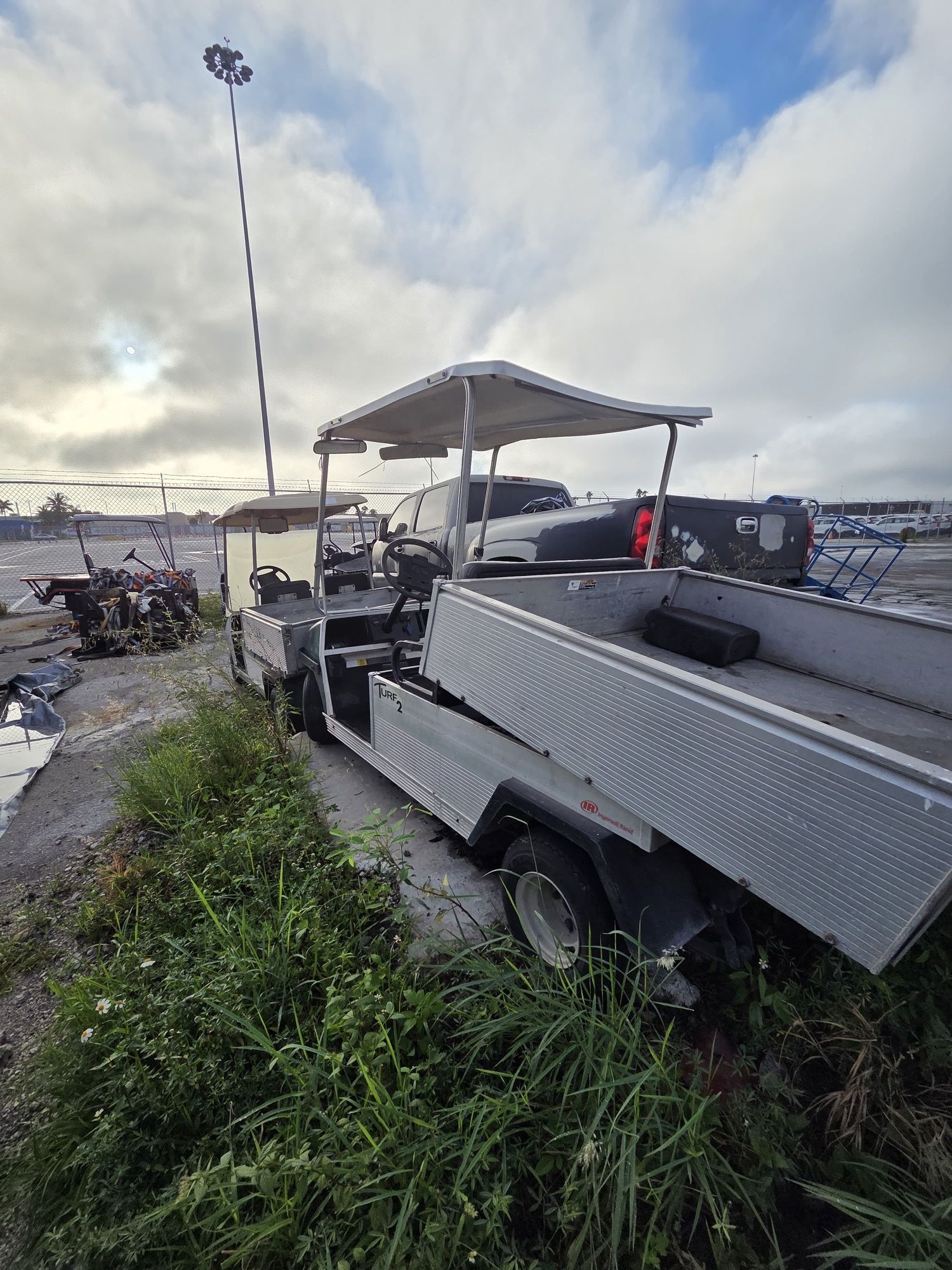 A silver utility vehicle with a canopy and trailer parked by tall grass under a cloudy sky.