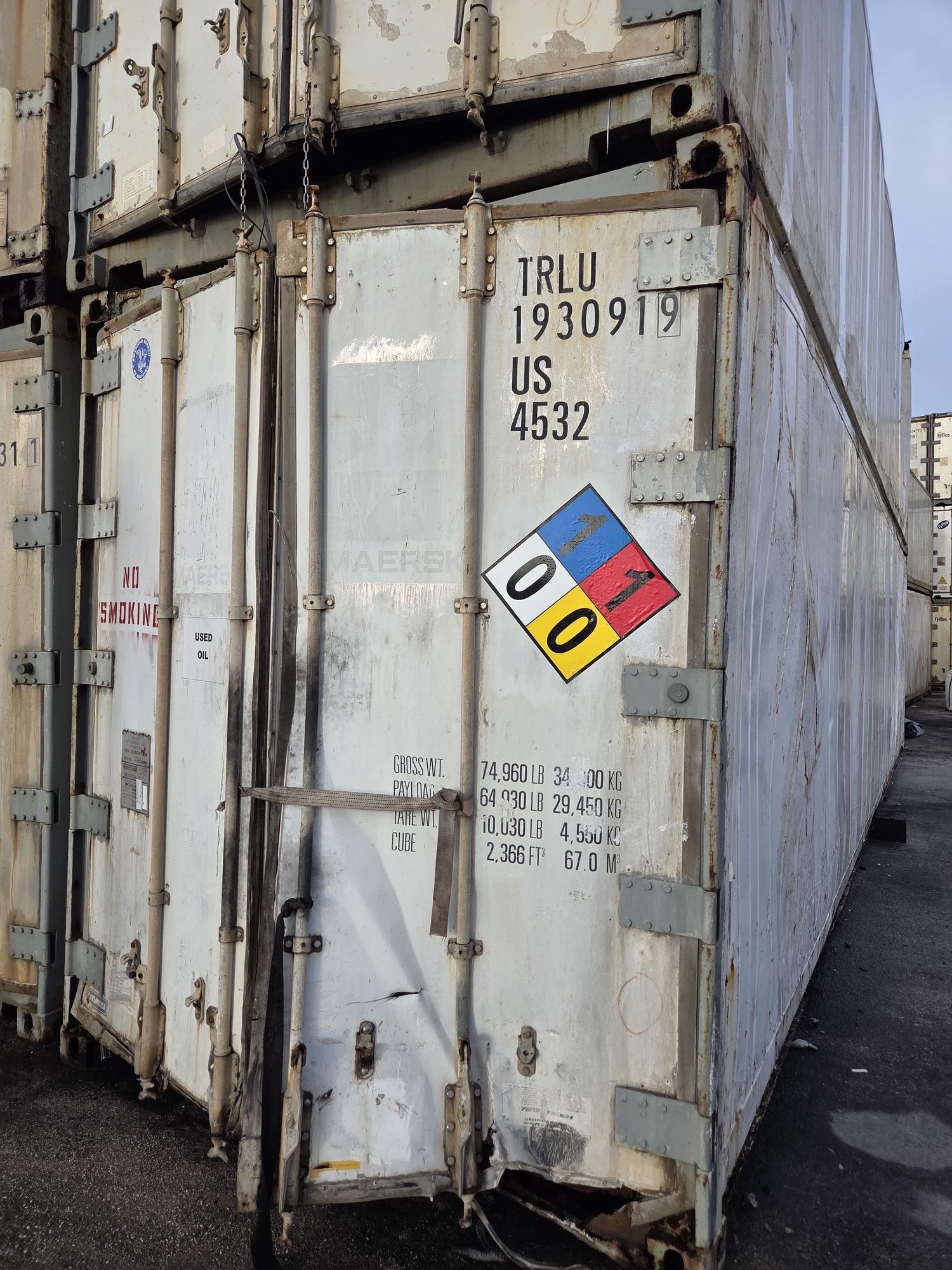 White shipping container with hazmat placard displaying a 00 code, stacked with other containers outside.