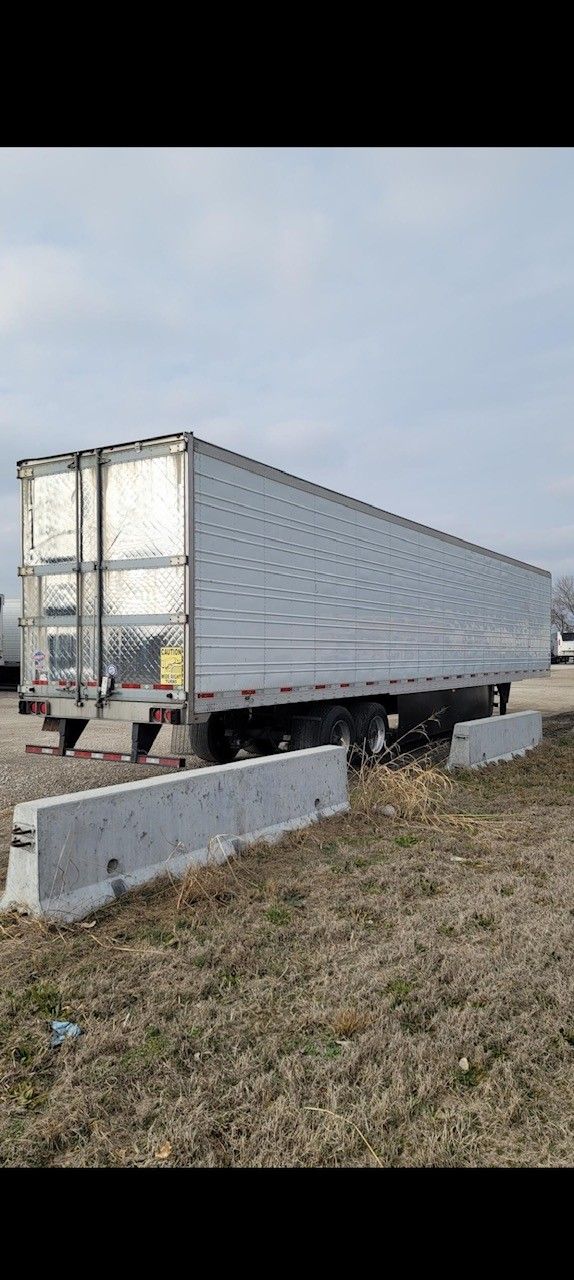 A large, empty silver semi-trailer parked on dry grass next to concrete barriers. Overcast sky.