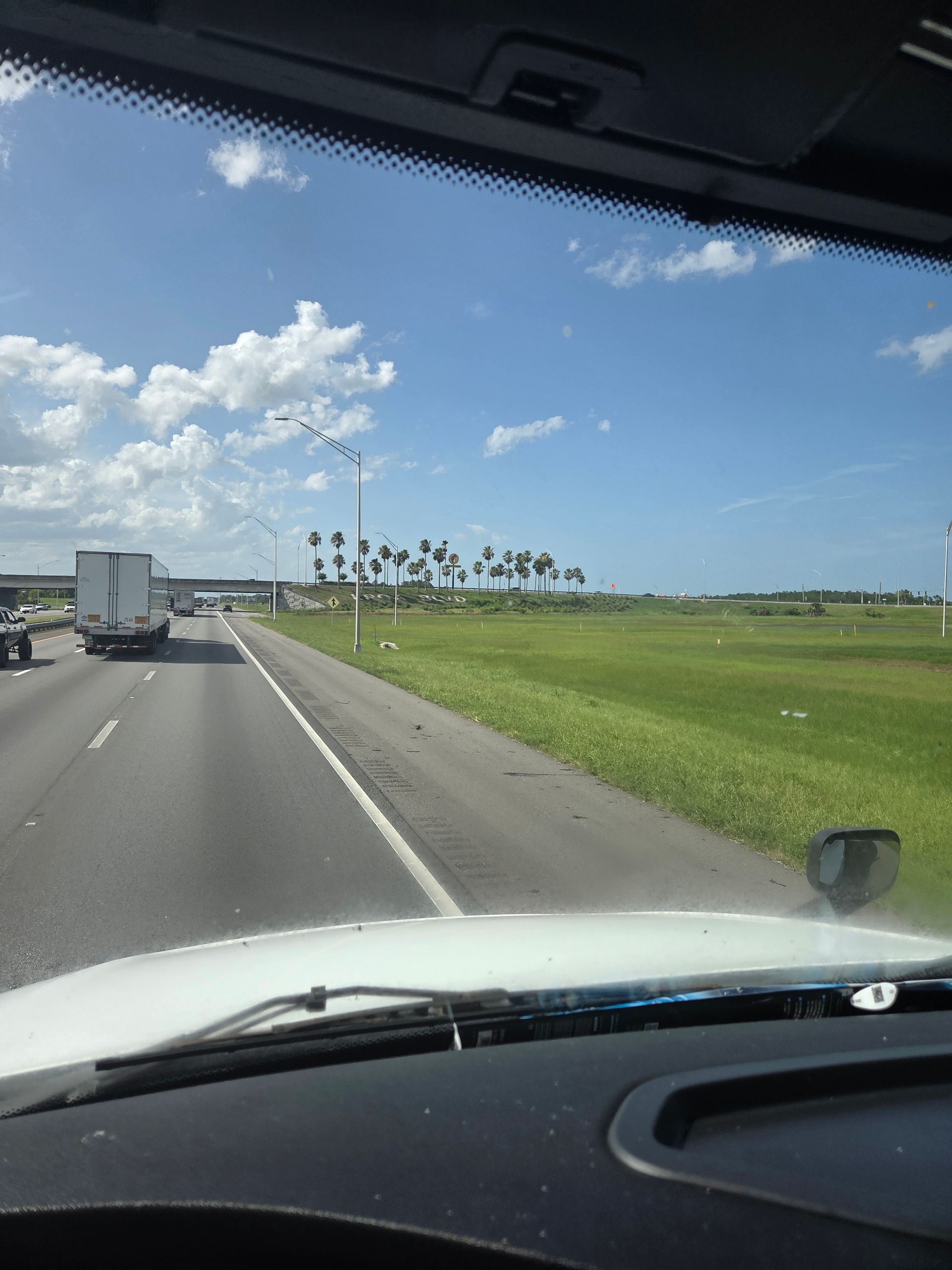 View from a vehicle's windshield on a sunny highway; semi-trucks on the road, grass field, and palm trees in the distance.
