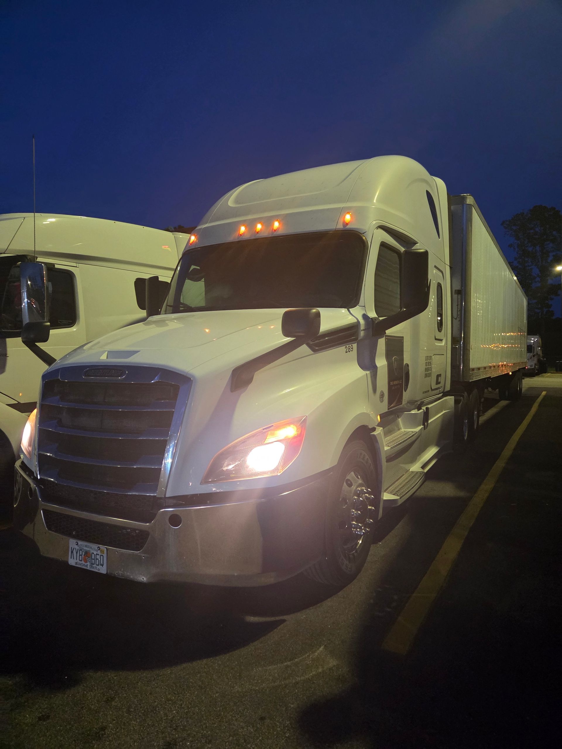White semi-truck with its headlights on, parked in a lot at dusk.