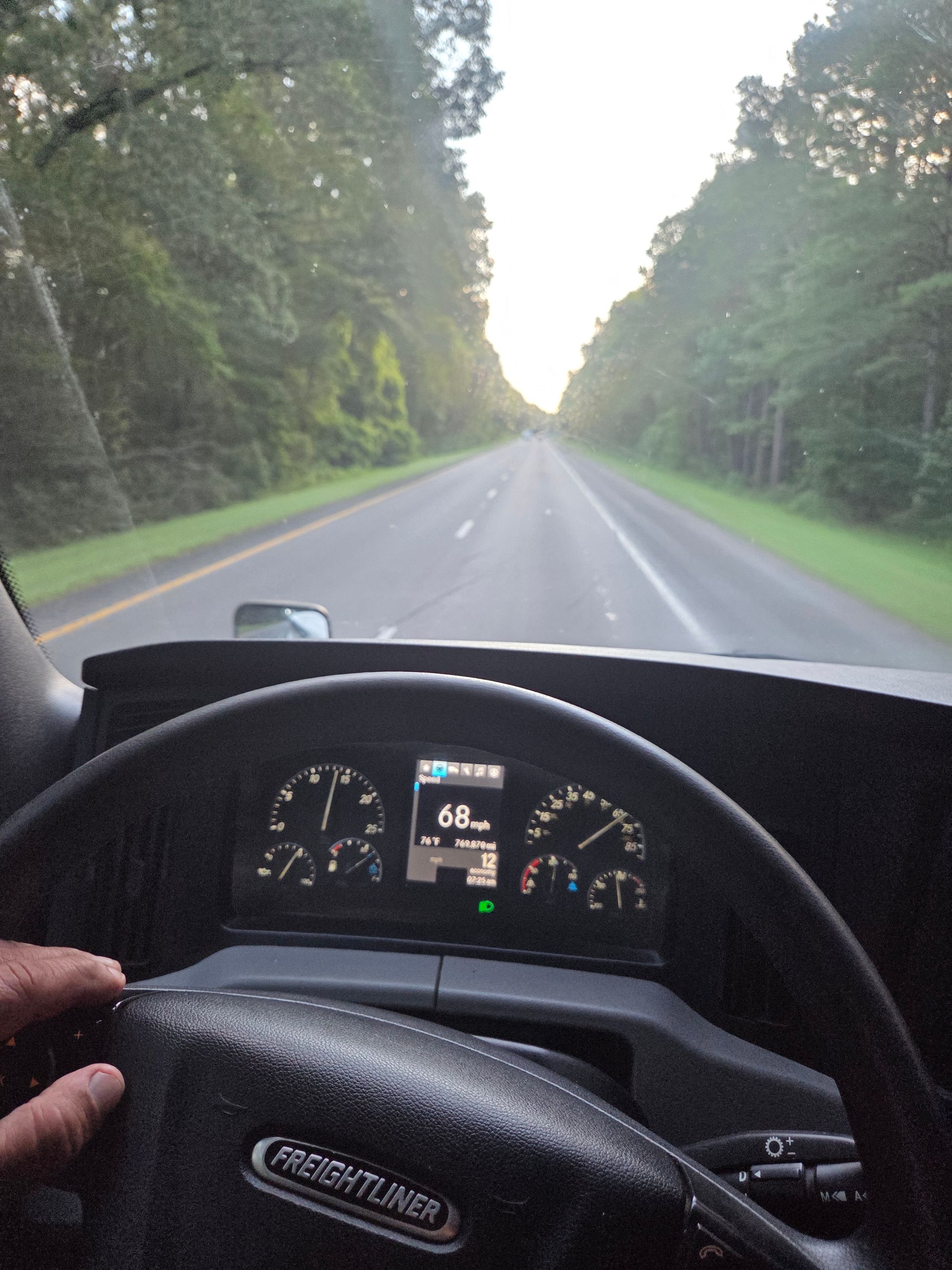 View from inside a Freightliner truck, driving on a highway, dashboard visible, trees in the background.