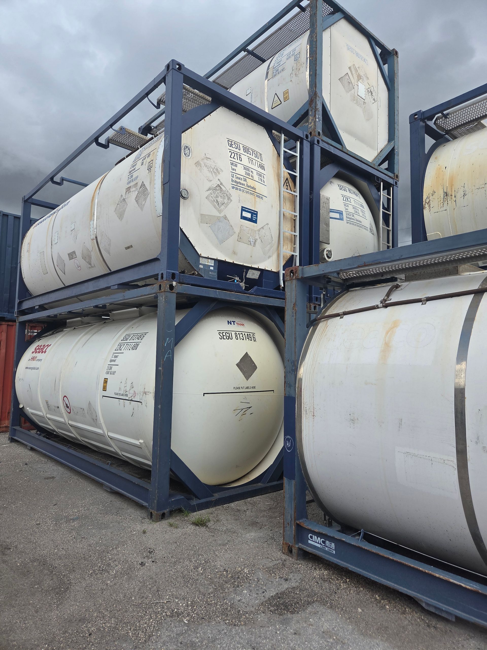 Stacked white tank containers in metal frames, outside on gravel.