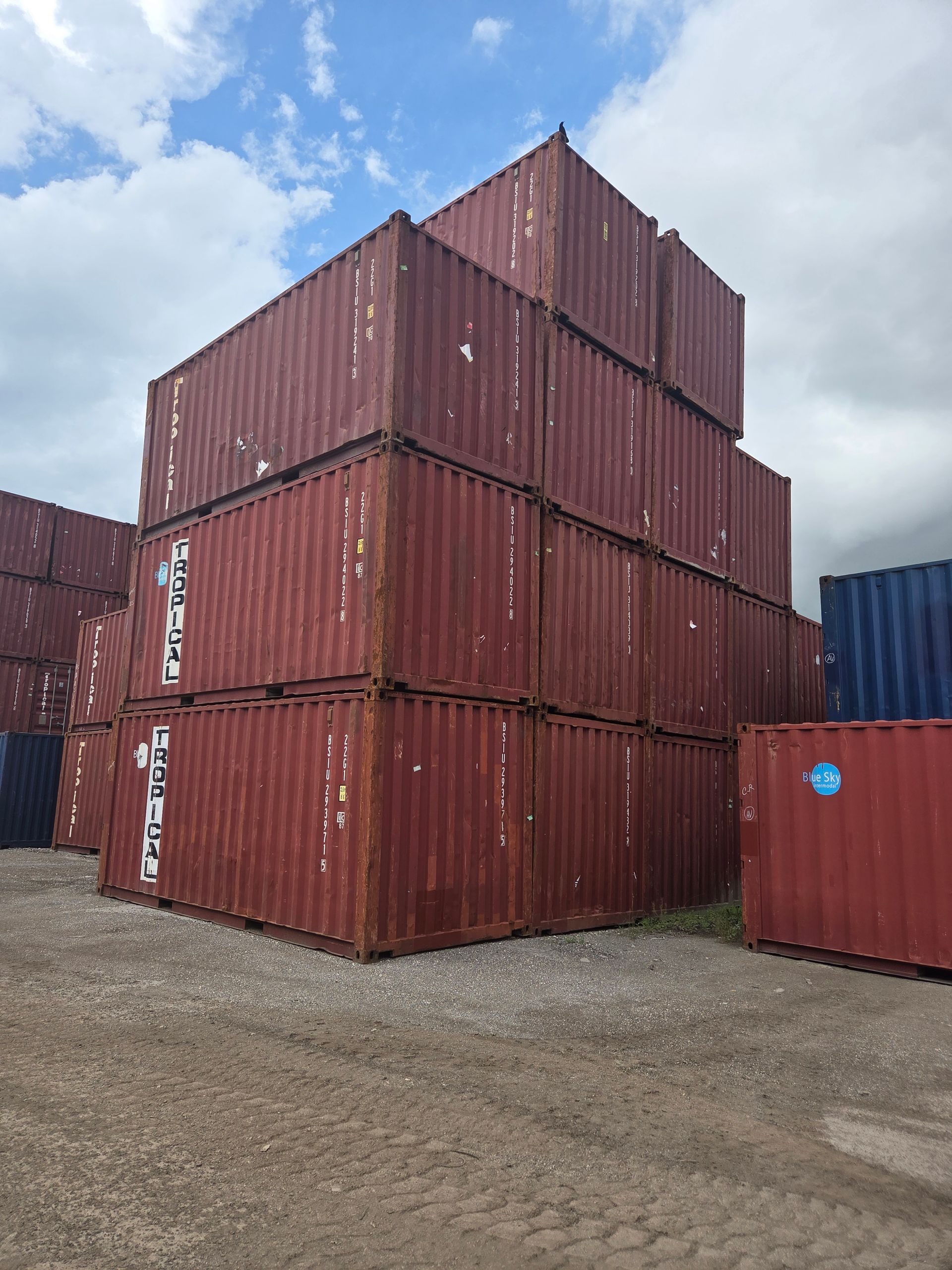 Stacked, rusty red shipping containers in an outdoor storage yard under a cloudy sky.