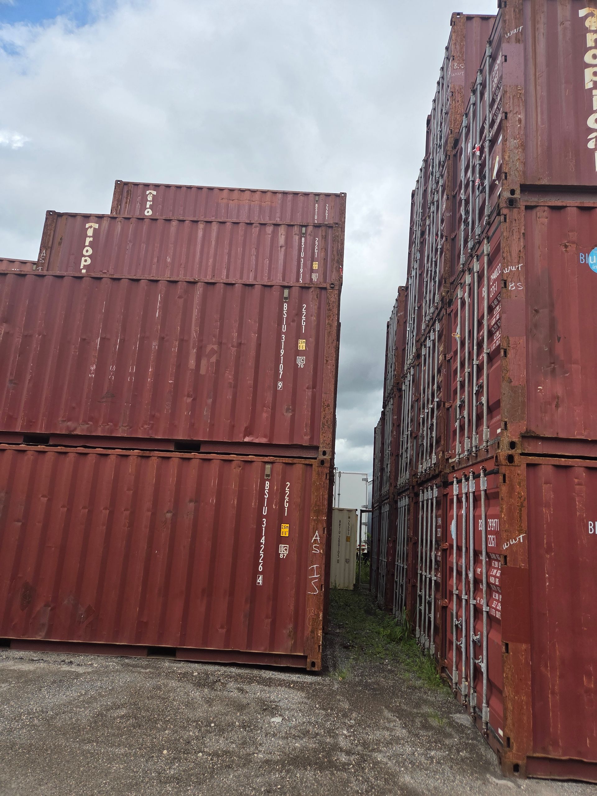 Red shipping containers stacked high outdoors, creating narrow passage. Cloudy sky overhead.