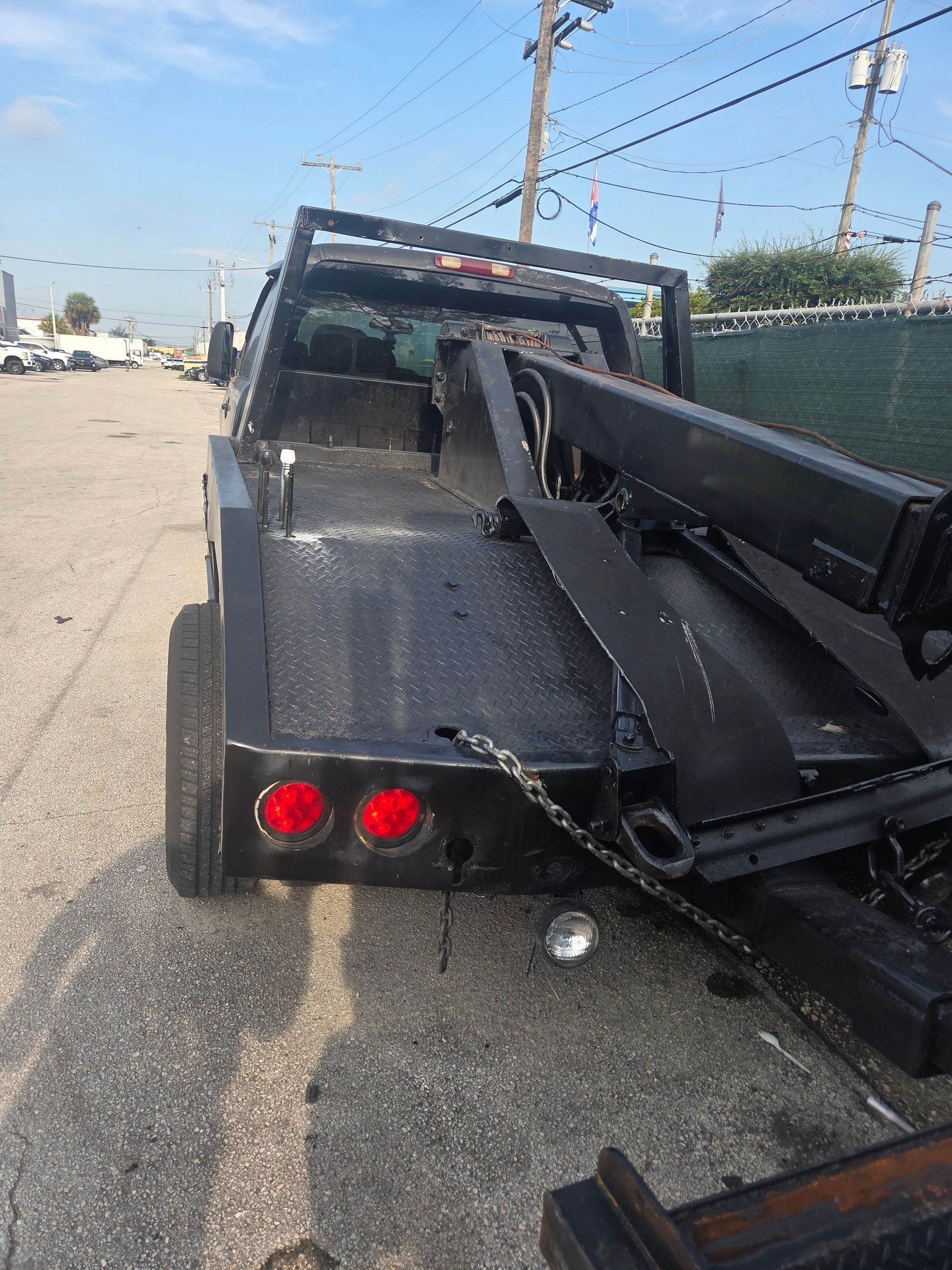 Black tow truck on a paved road, daytime. Rear view, with red tail lights, tow arm extended.