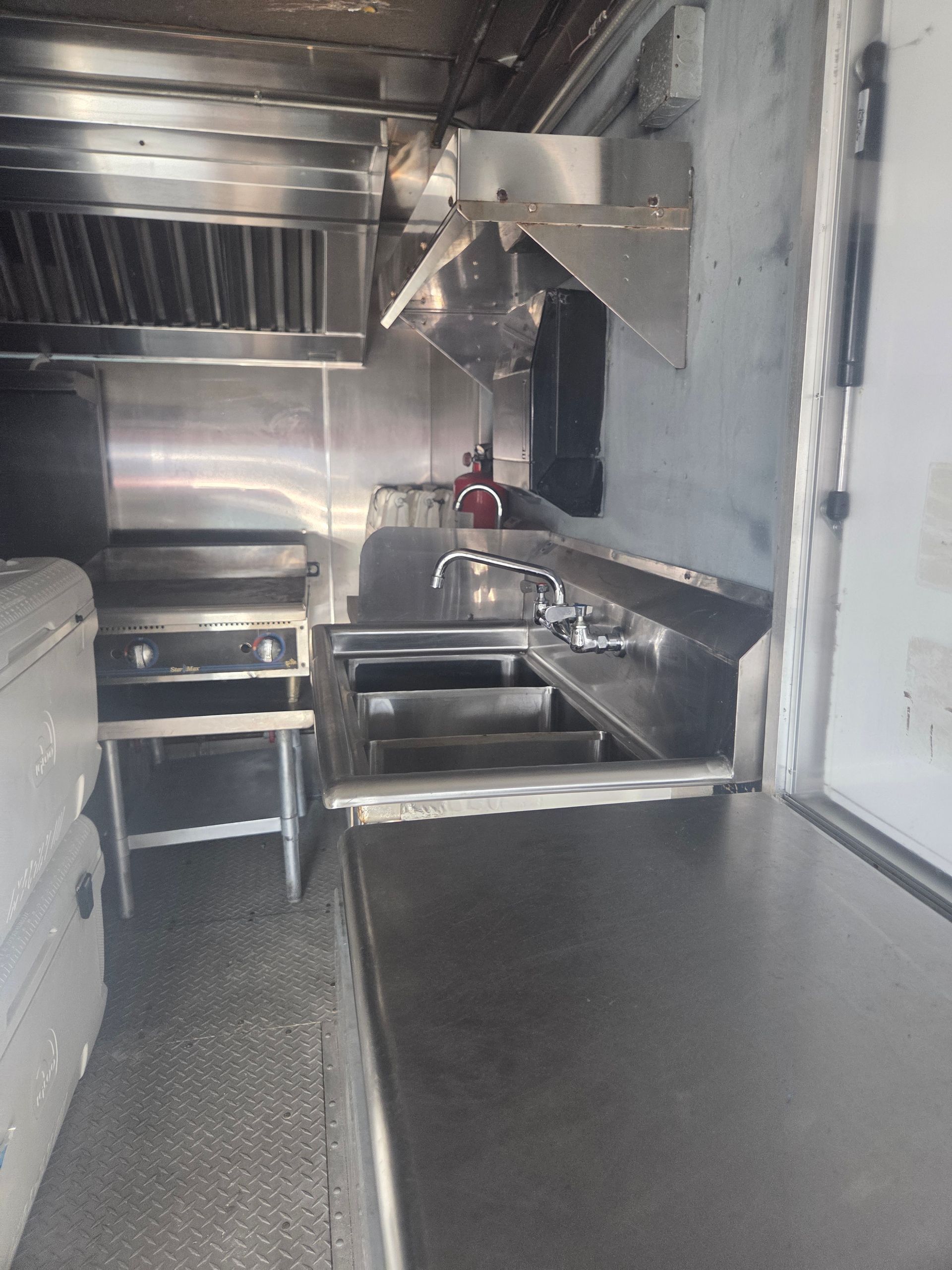 Interior of a food truck kitchen: stainless steel appliances, sinks, and a hood with a grease fire extinguisher.