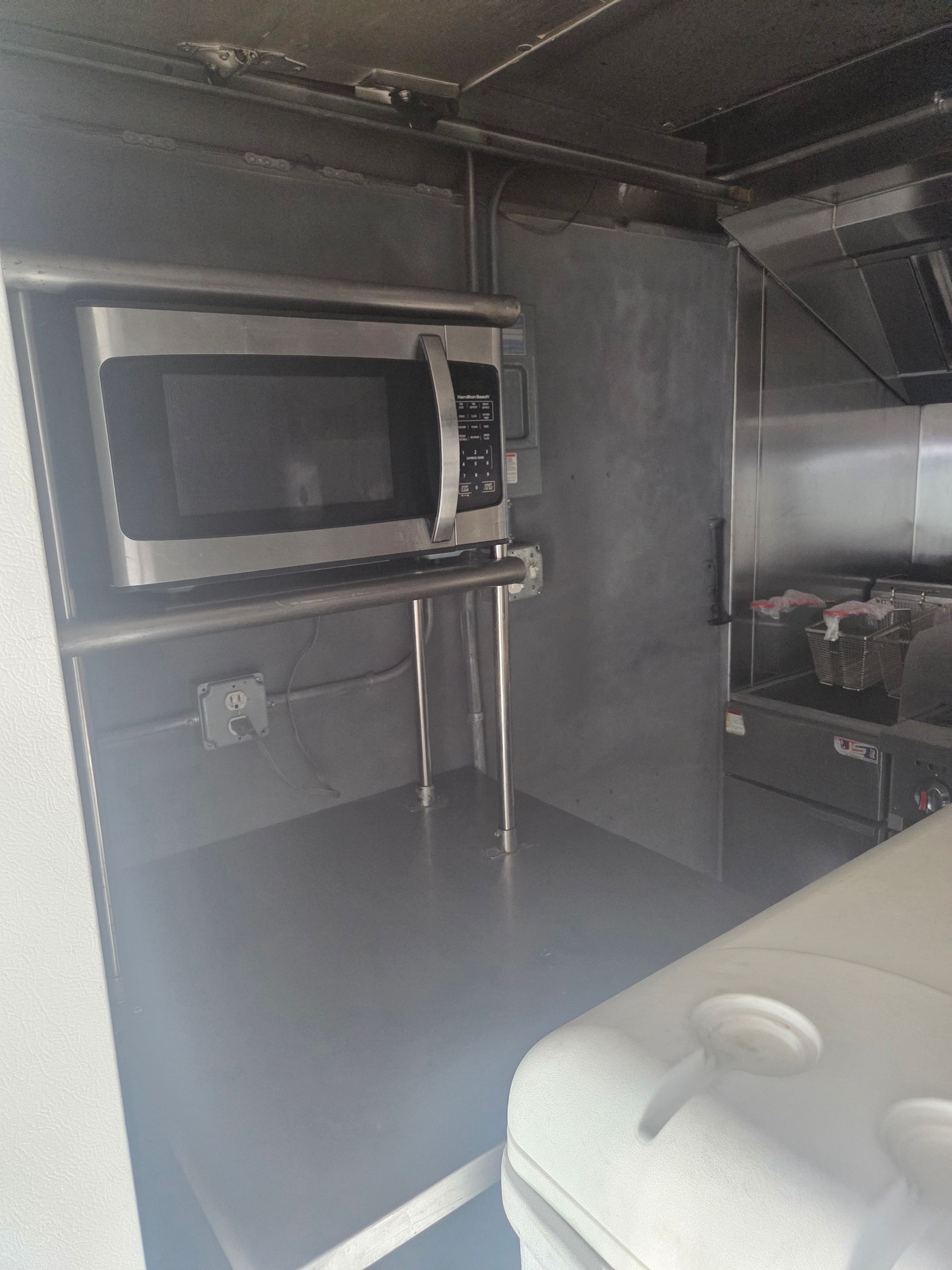 Stainless steel food truck interior with a microwave, shelf, and cooking equipment.