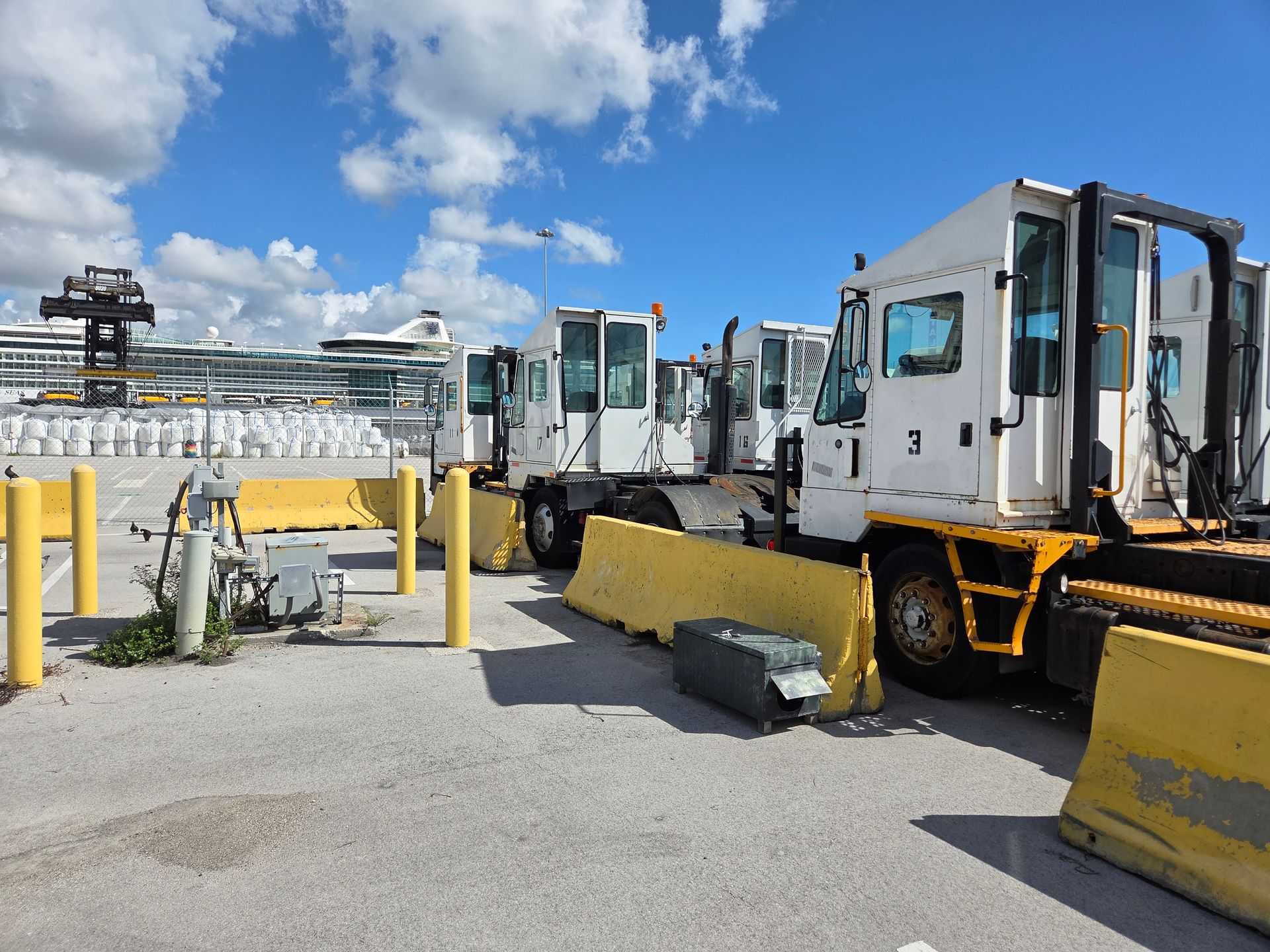 White terminal tractors parked behind yellow barriers at a port, cruise ships in the background under a blue sky.