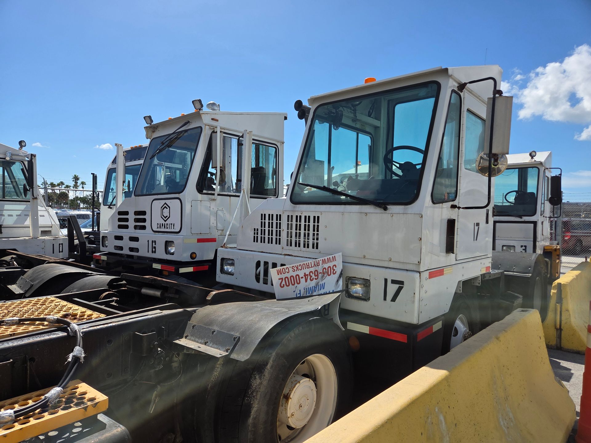 Several white terminal tractors parked in a lot, sunny day, blue sky.