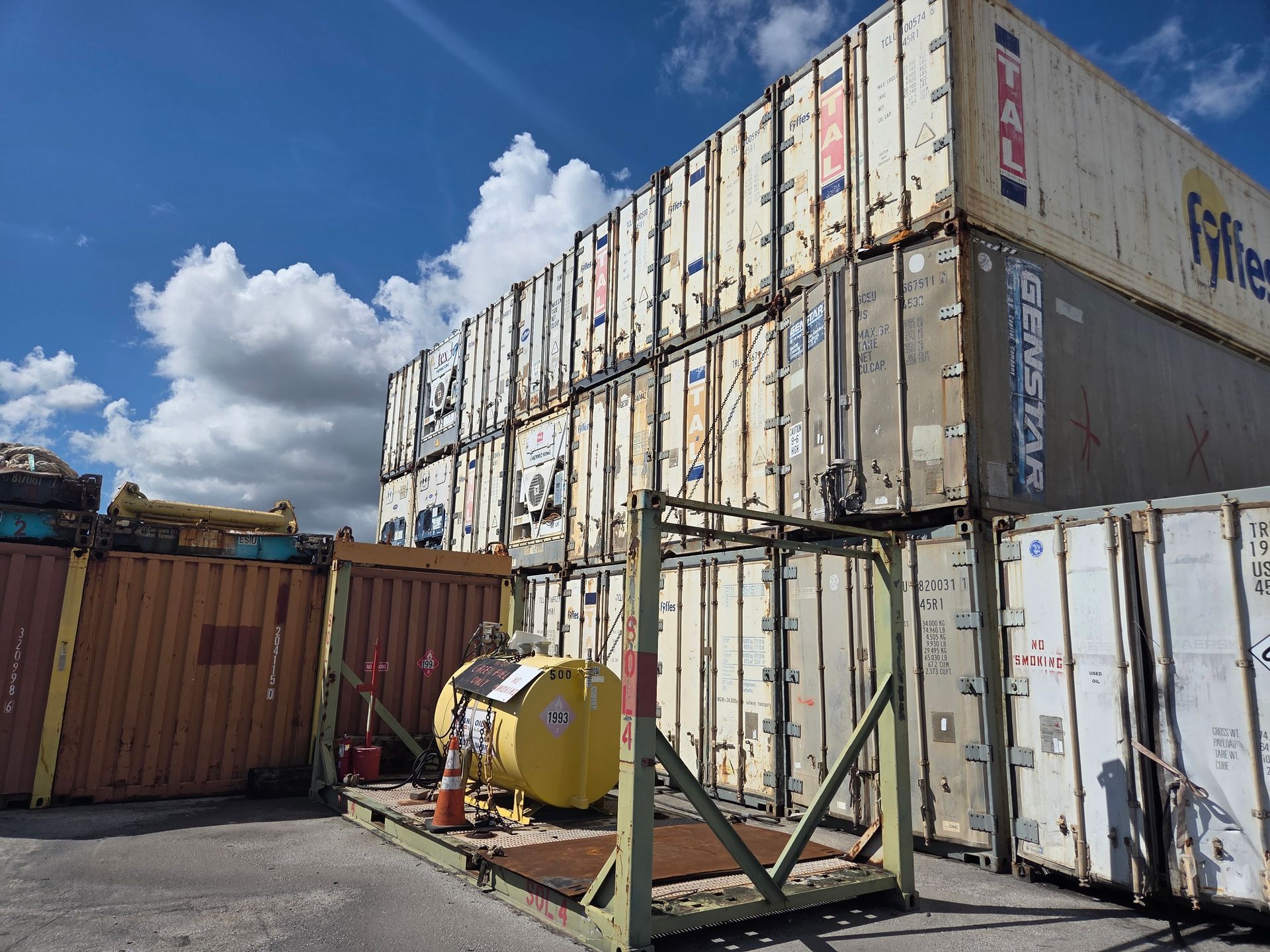 Stacked shipping containers with a yellow tank on a metal stand, under a blue sky.