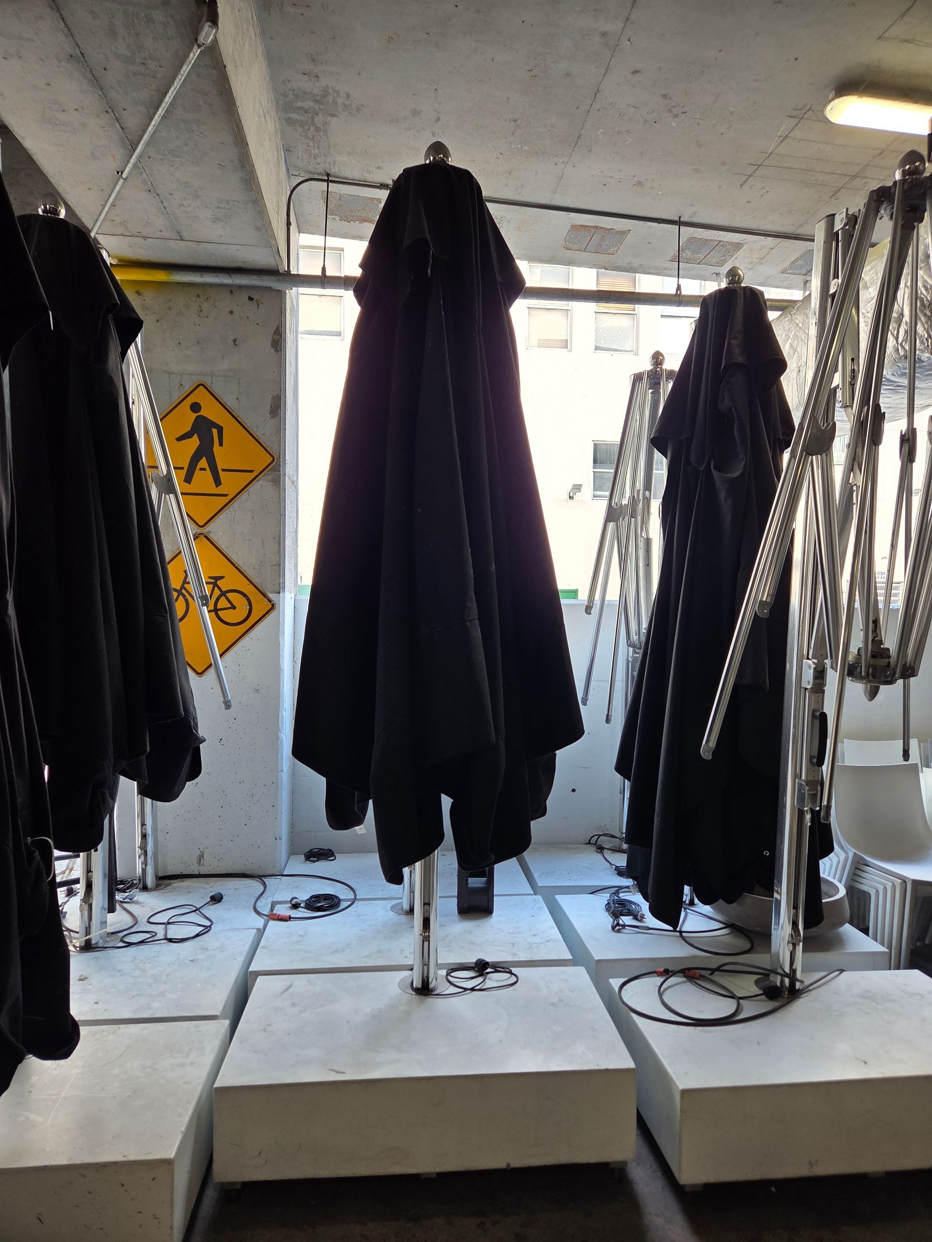 Closed black umbrellas on metal stands, in a concrete room. Pedestrian and bike crossing sign visible.