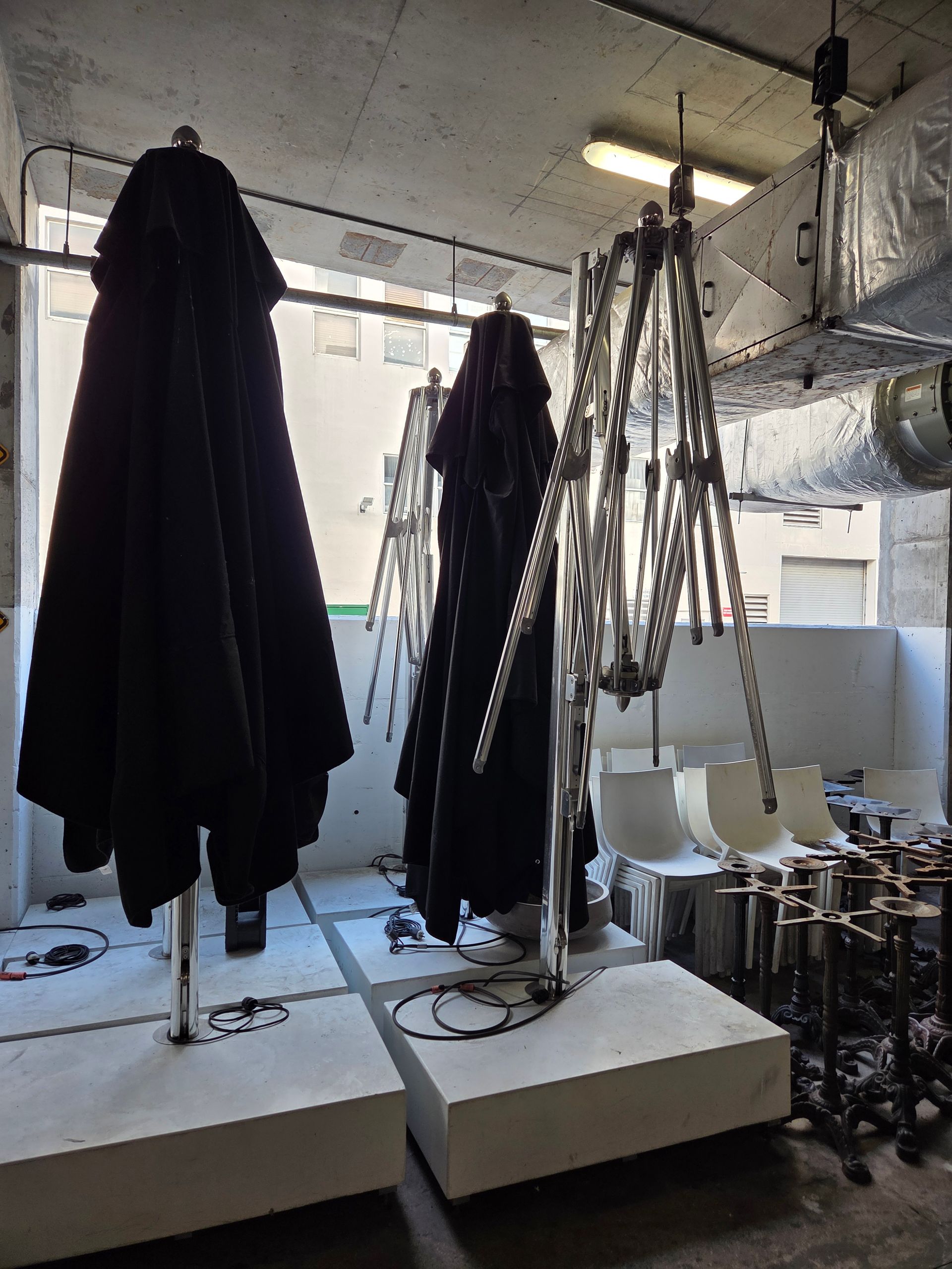 Three black patio umbrellas folded, on concrete bases, indoors. Stainless steel frames, white walls.