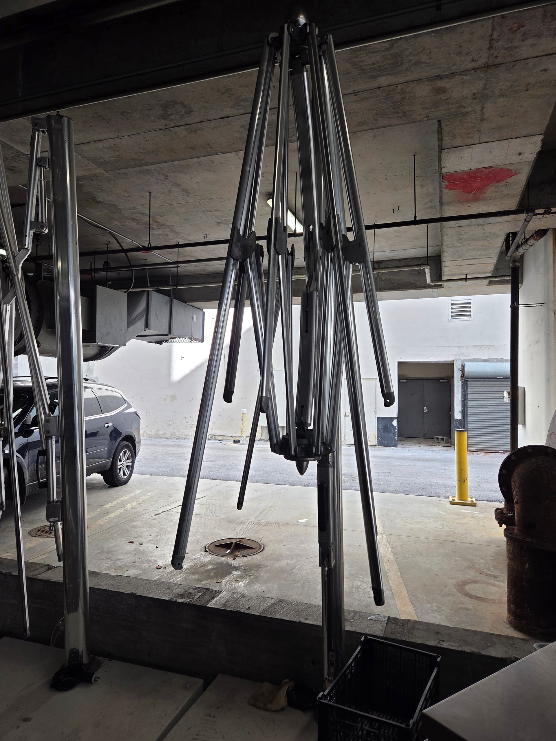 Metal frame suspended from a ceiling, potentially for hanging objects, in an open-air area with a parked vehicle.