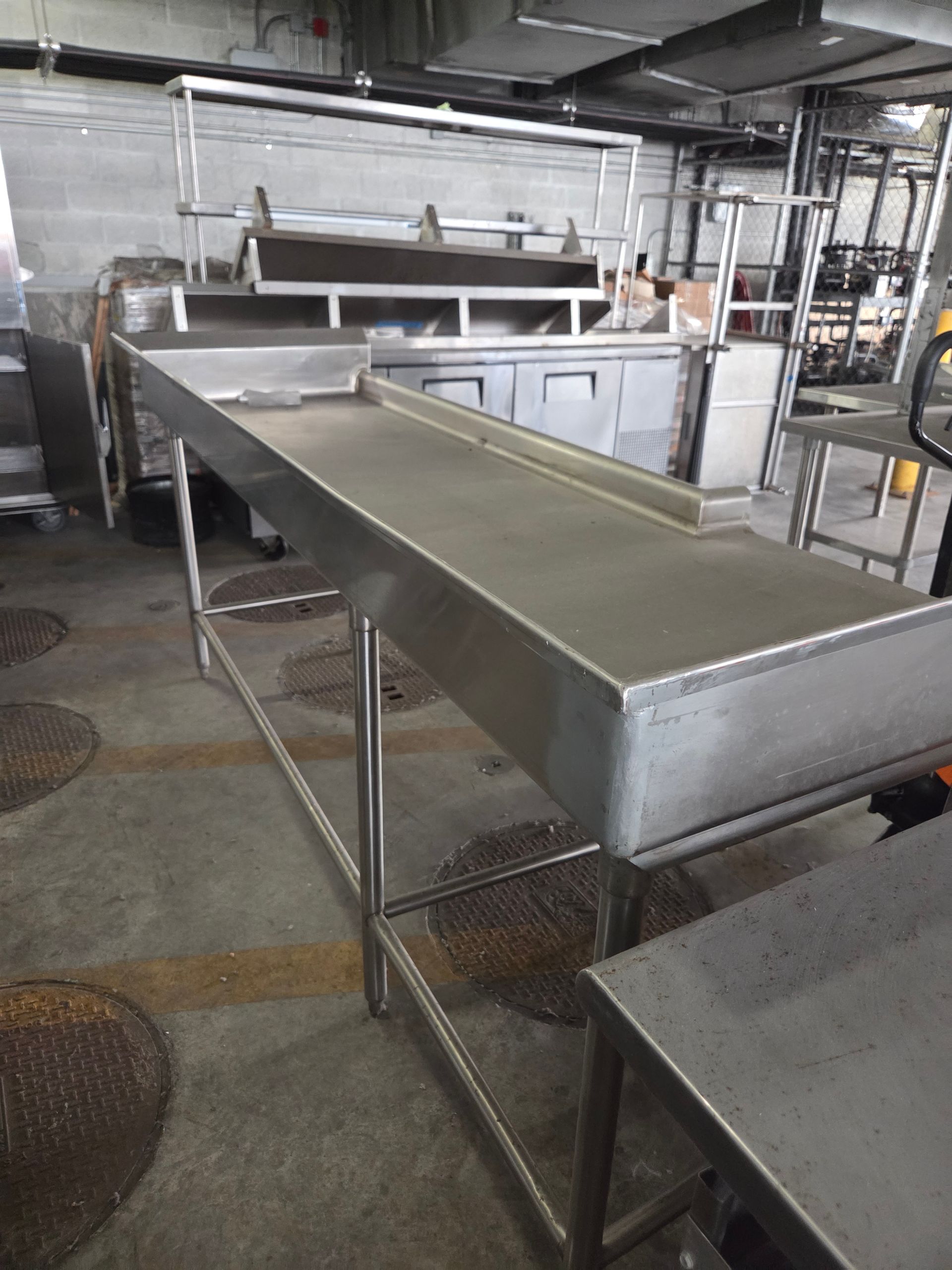 Stainless steel work table in a commercial kitchen with other equipment visible.