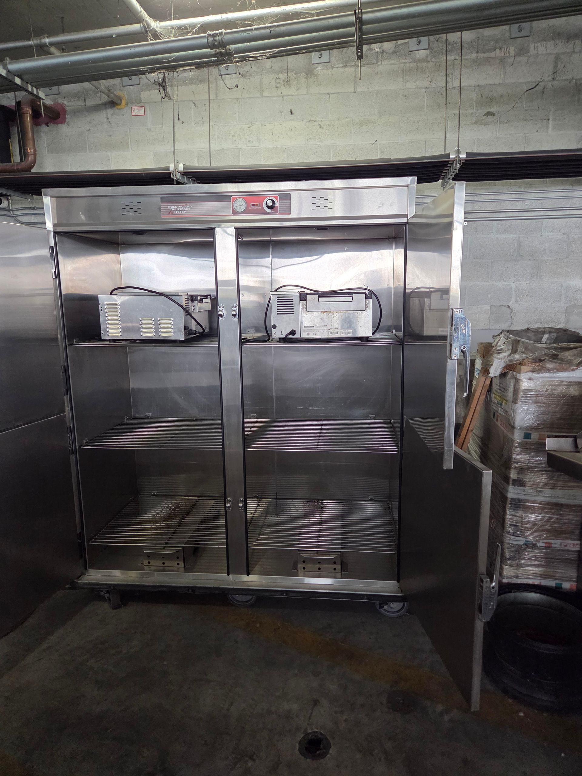 Stainless steel refrigerator with shelves holding equipment, in a concrete-walled room.