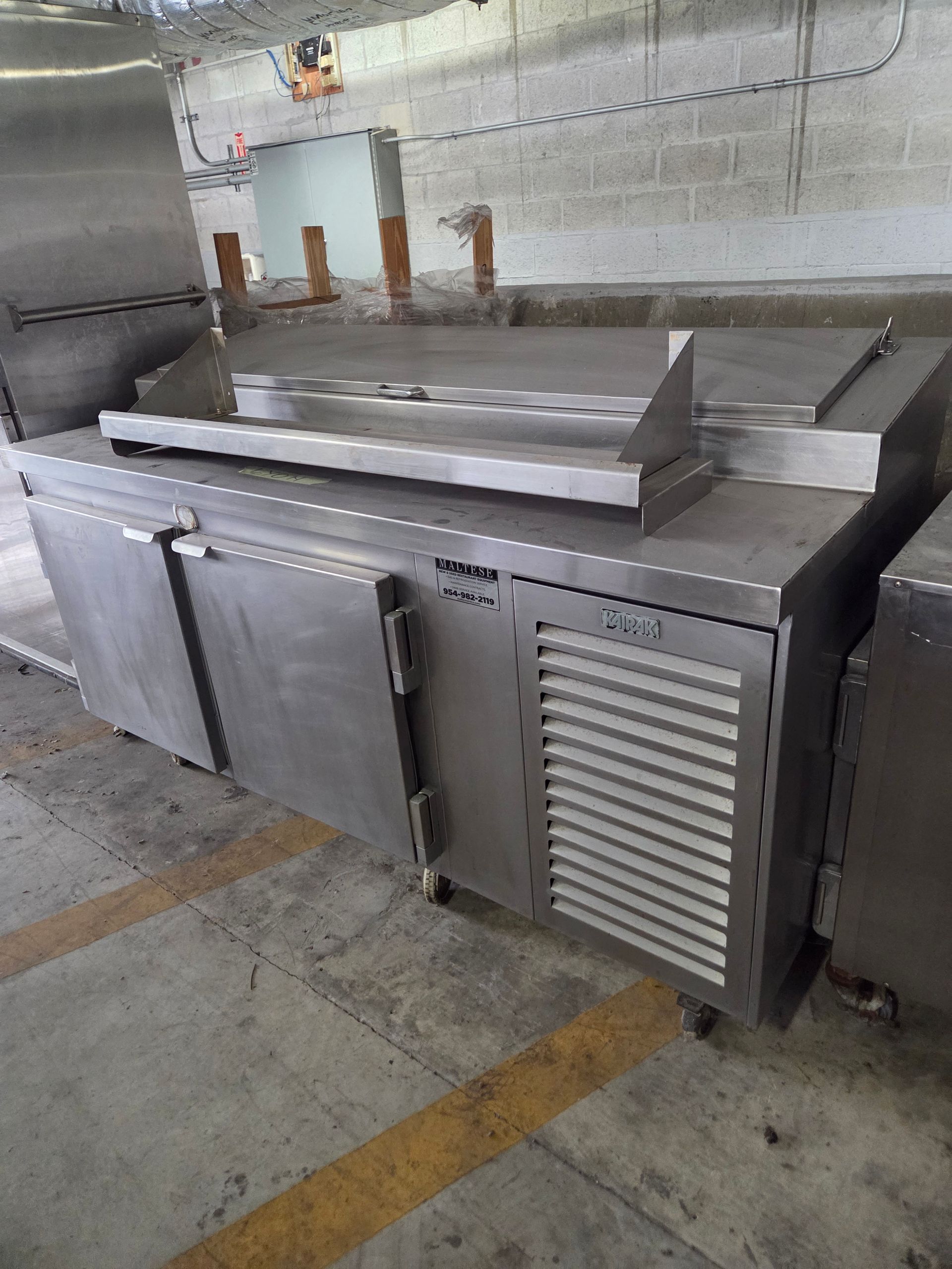 Stainless steel commercial kitchen refrigeration unit with two doors, a vent, and a countertop, in a warehouse setting.