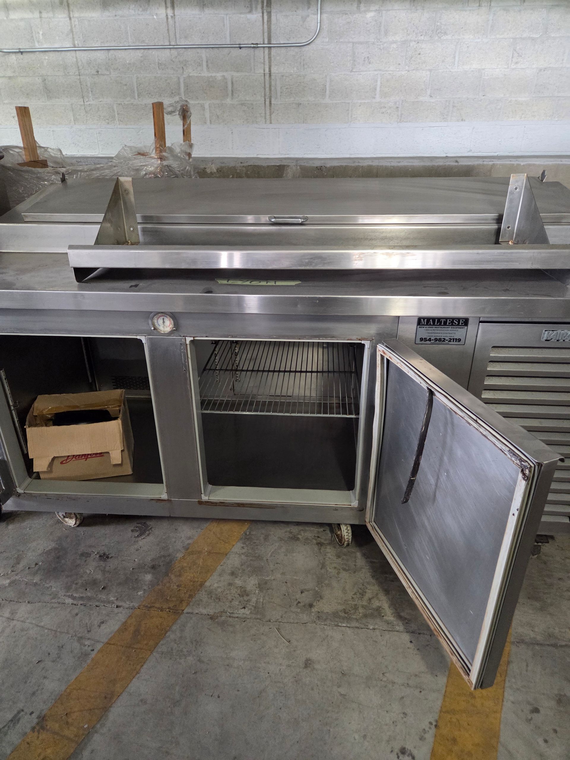 Stainless steel commercial refrigerator with an open door, revealing shelves. Located in a concrete setting.