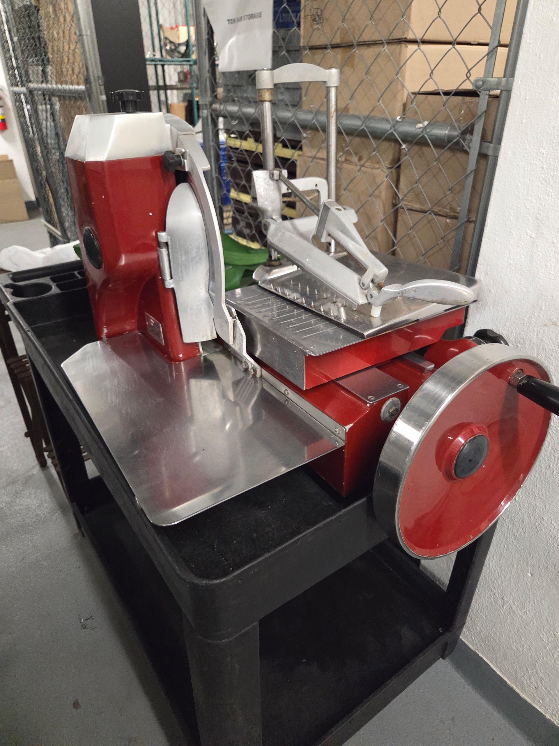 Red commercial meat slicer on a black cart in a storage area.