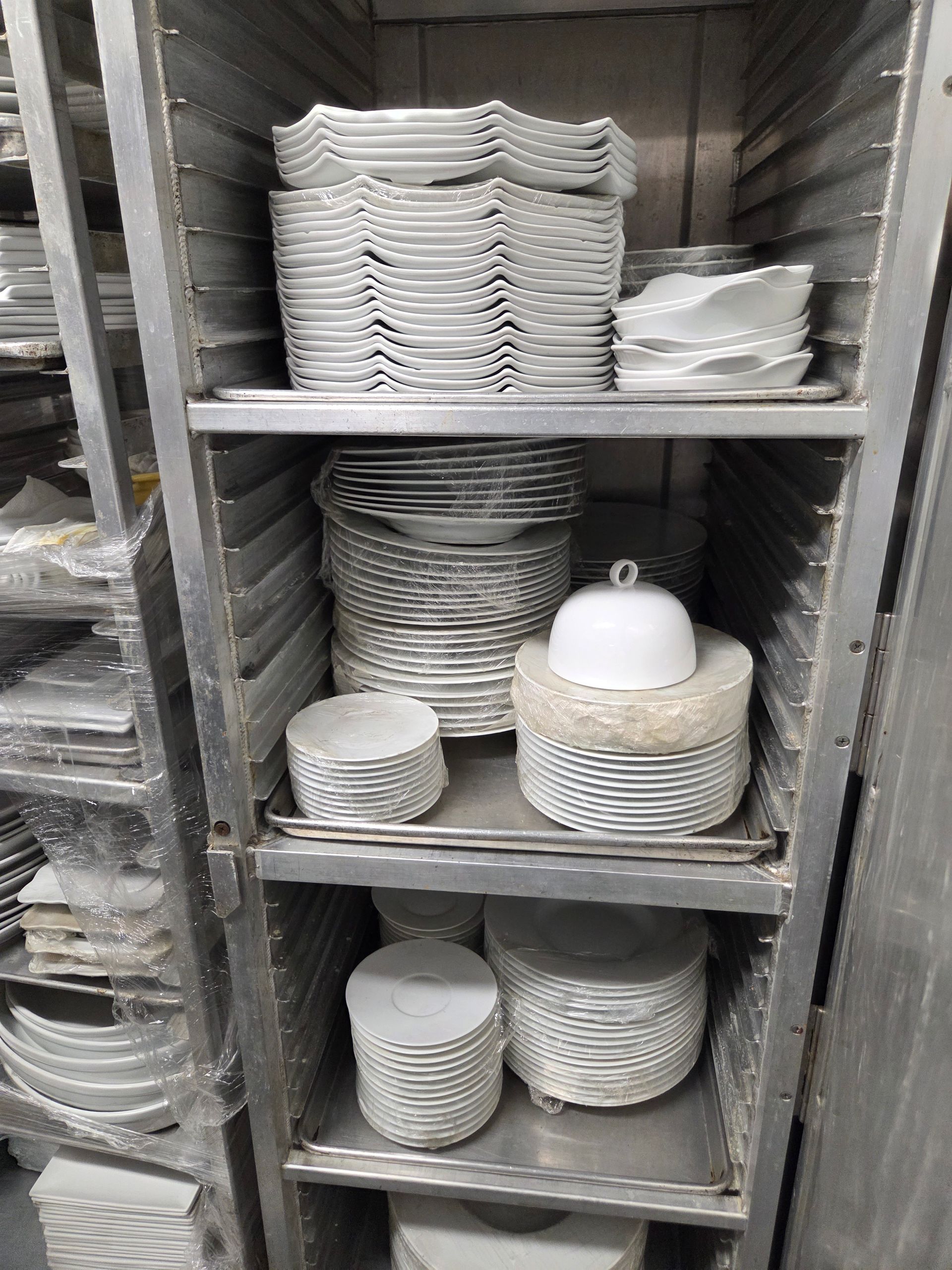 Shelves holding stacks of white plates, bowls, and a covered dish in a commercial kitchen storage unit.