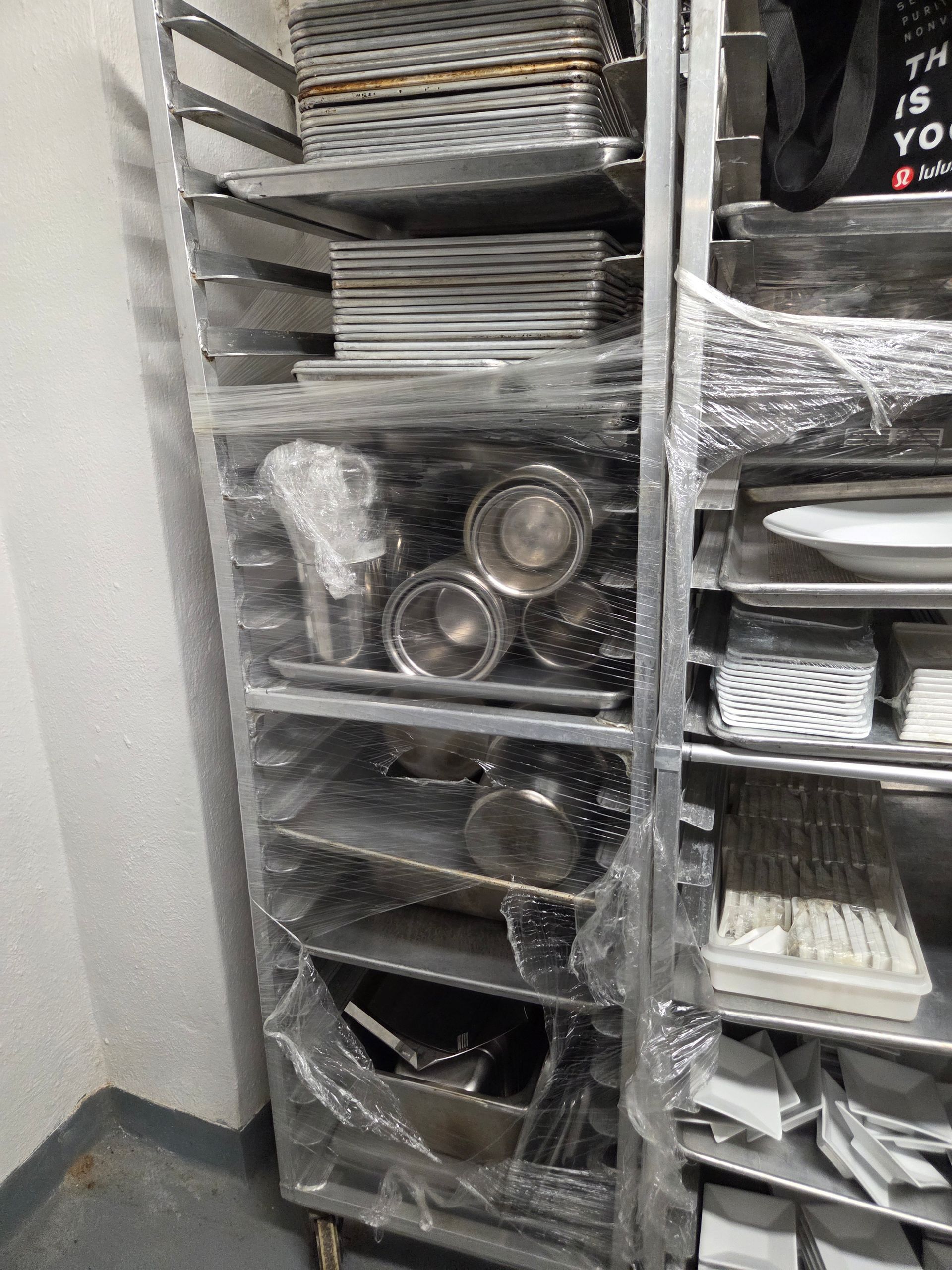 Metal shelving unit holding various kitchen dishes and cookware, wrapped in plastic.