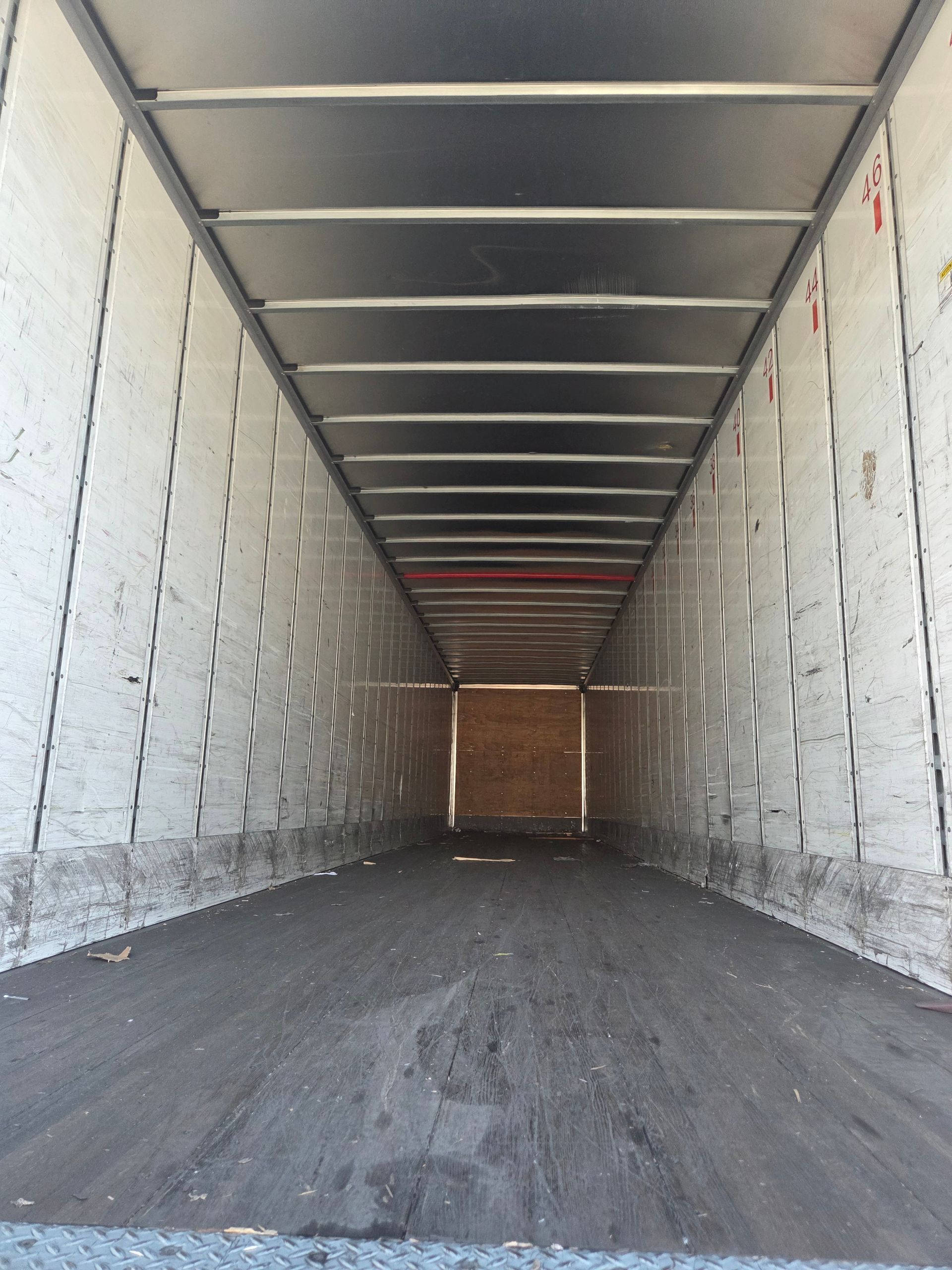 Interior of an empty semi-truck trailer, with metal roof and walls, and dark flooring extending to a closed door.