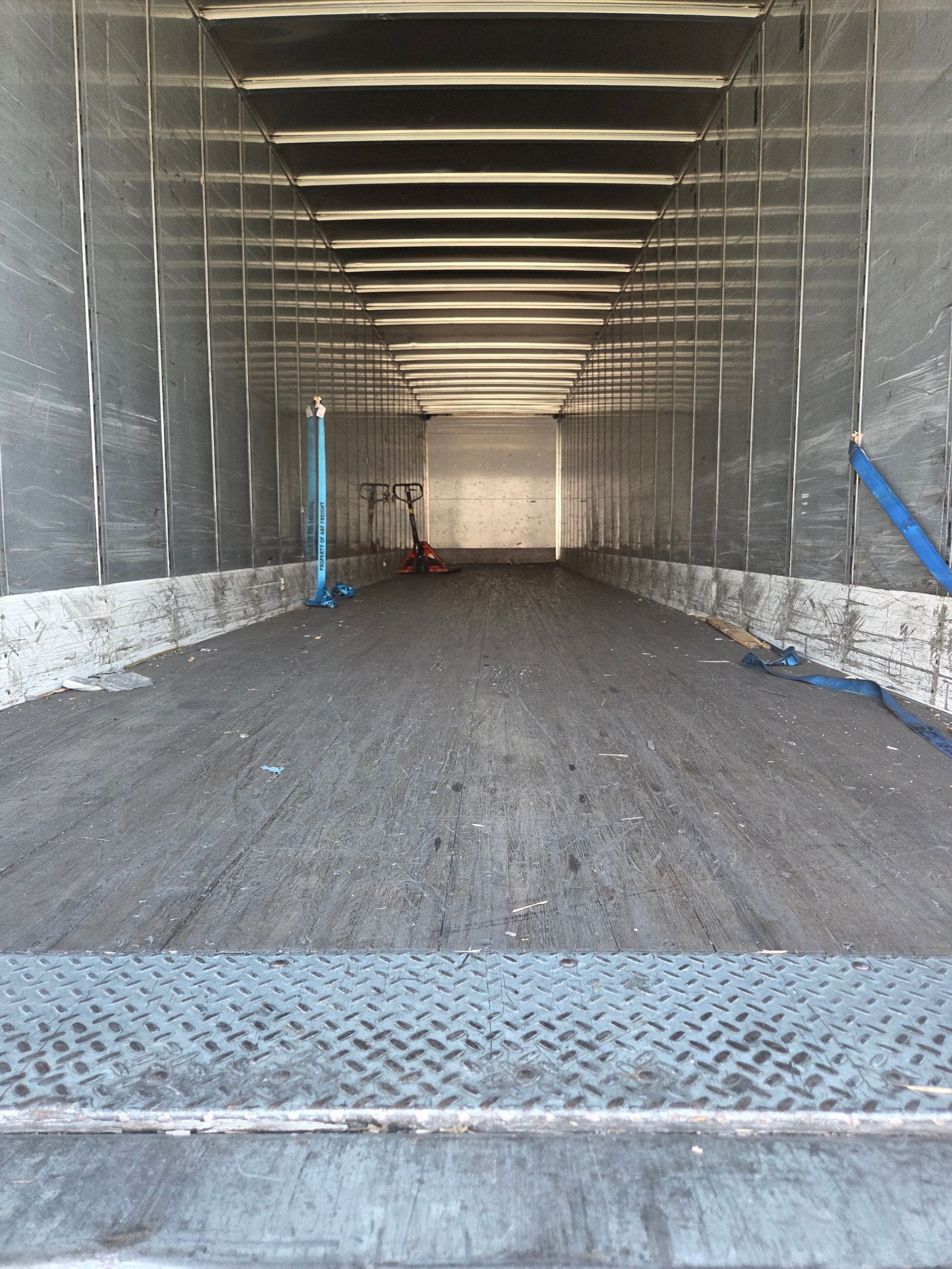 Interior of an empty semi-truck cargo area. Gray floor and walls, metal frame. Back open to sunlight.