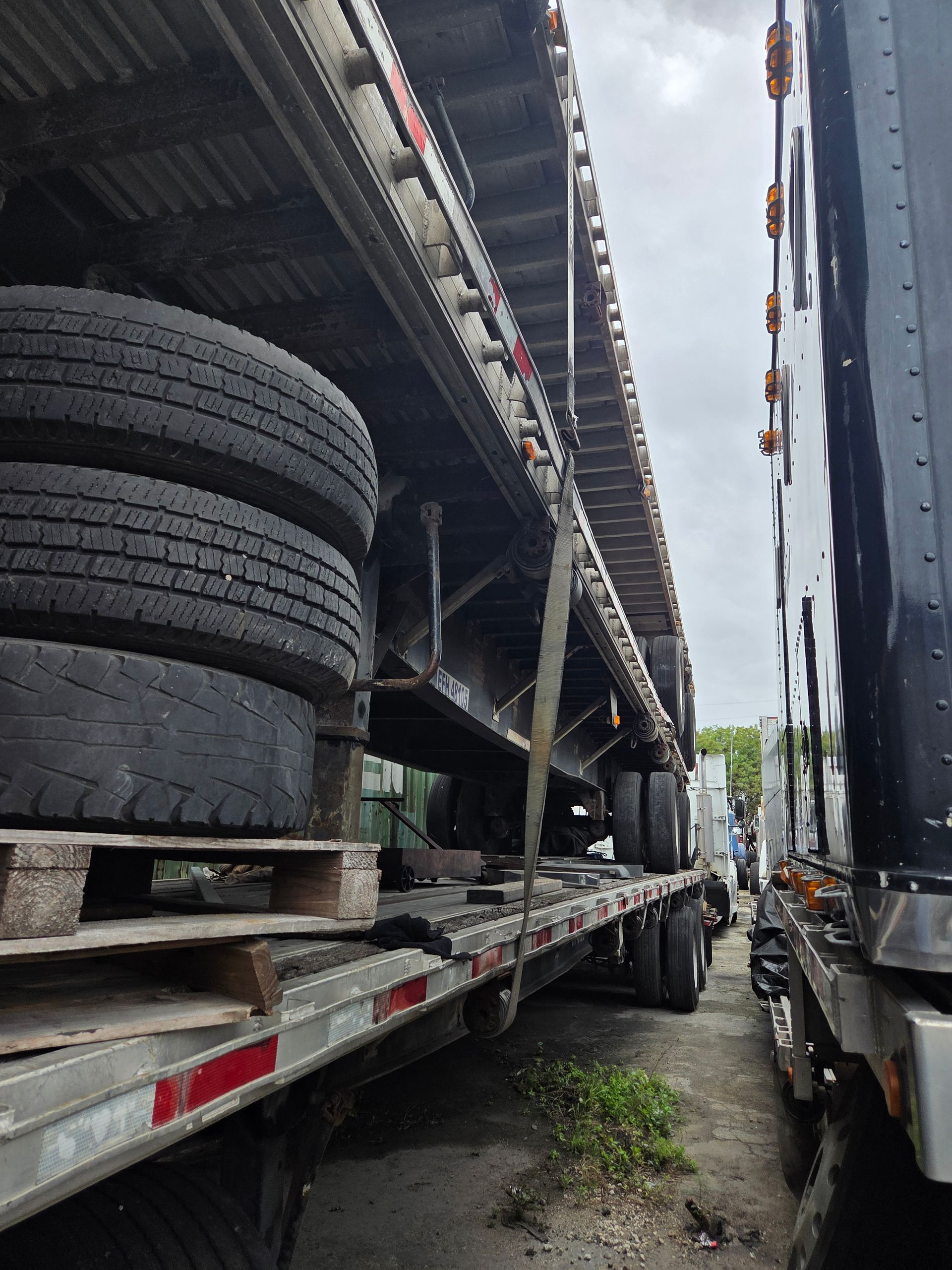 Flatbed trailer loaded with tires and a truck, parked on asphalt. Overcast sky.