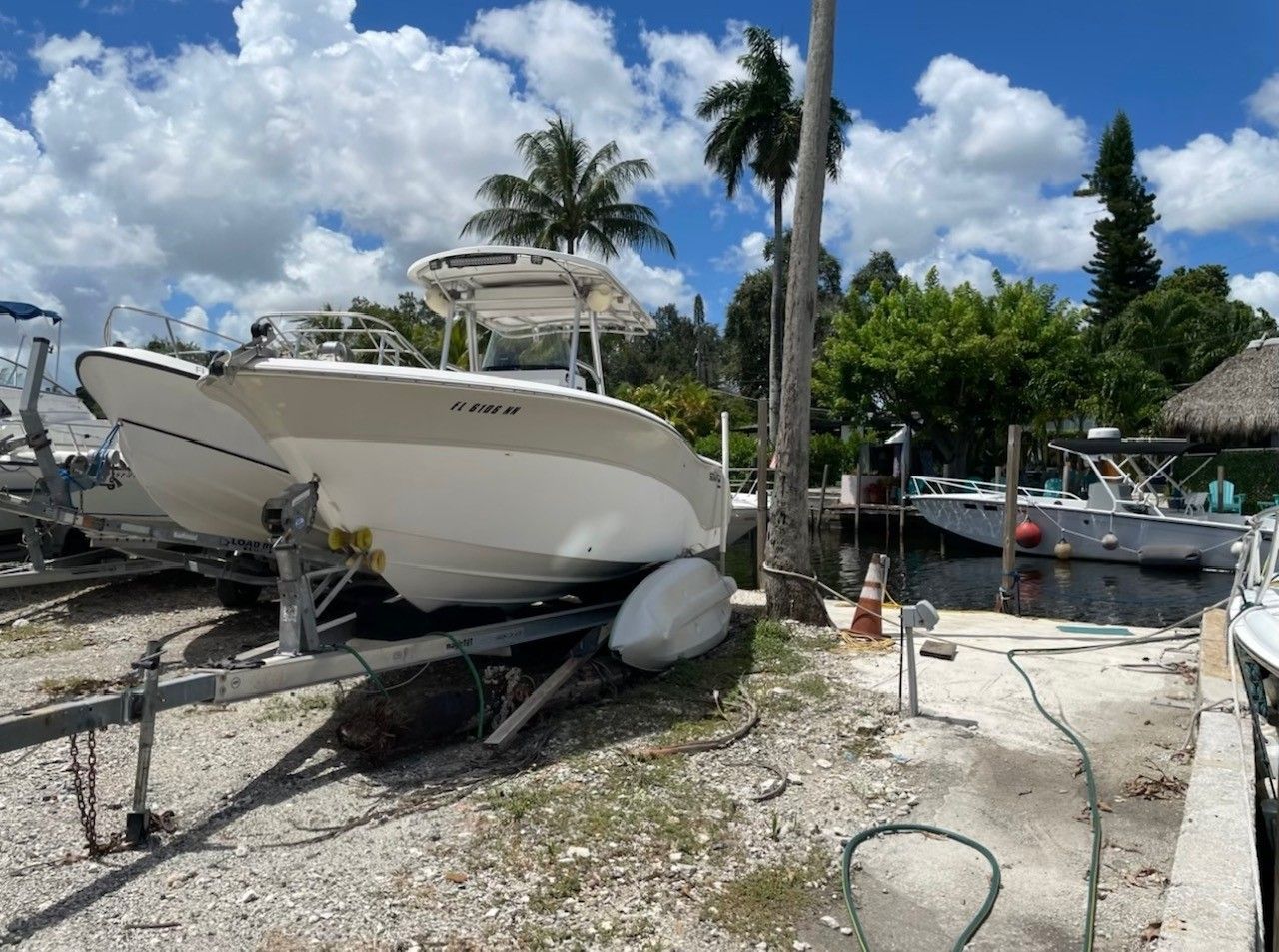 A white boat on a trailer parked on a gravel surface near a body of water with palm trees in the background.