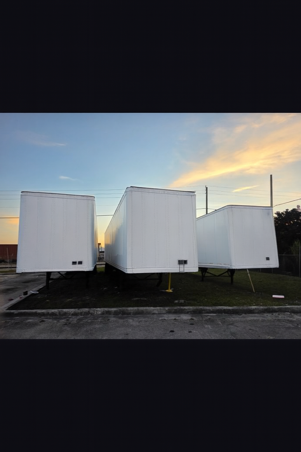 Three white semi-trailer trucks parked outdoors on a grassy area, against a blue and yellow sunset sky.
