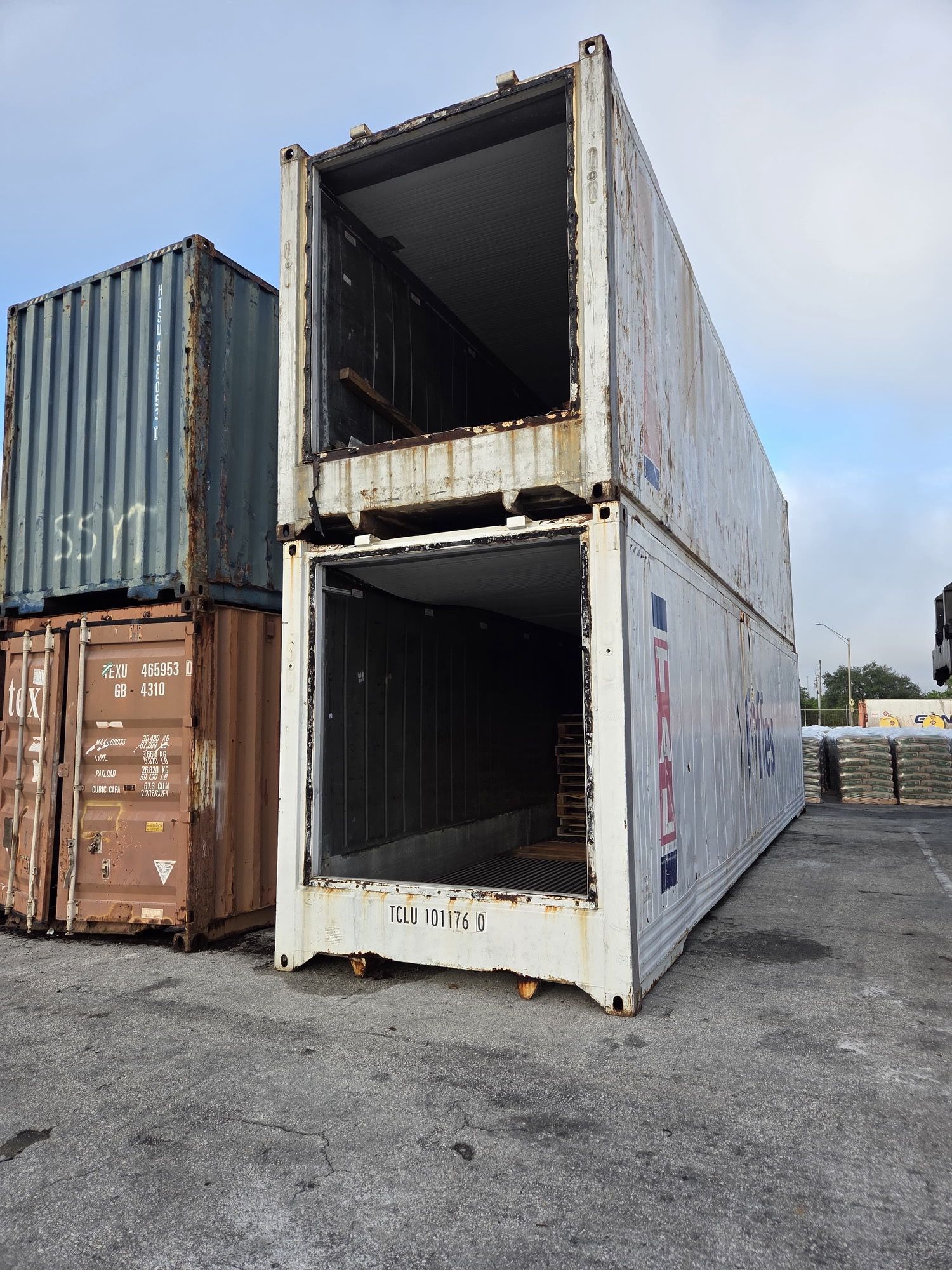 Two stacked, open-ended shipping containers in a storage yard; weathered, white container on top, blue and brown ones behind.