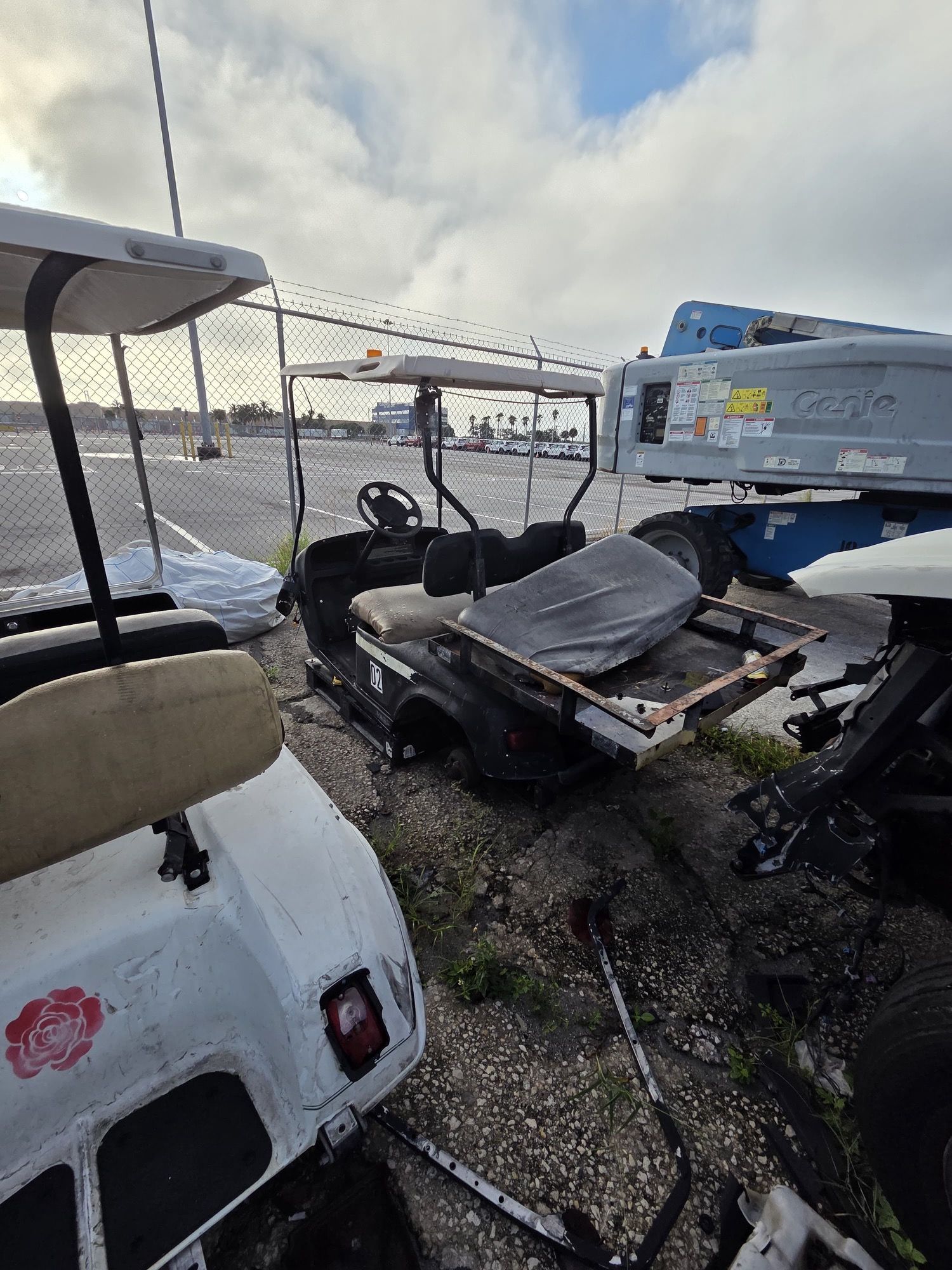 Golf carts parked outdoors near a fence and machinery, under a cloudy sky.