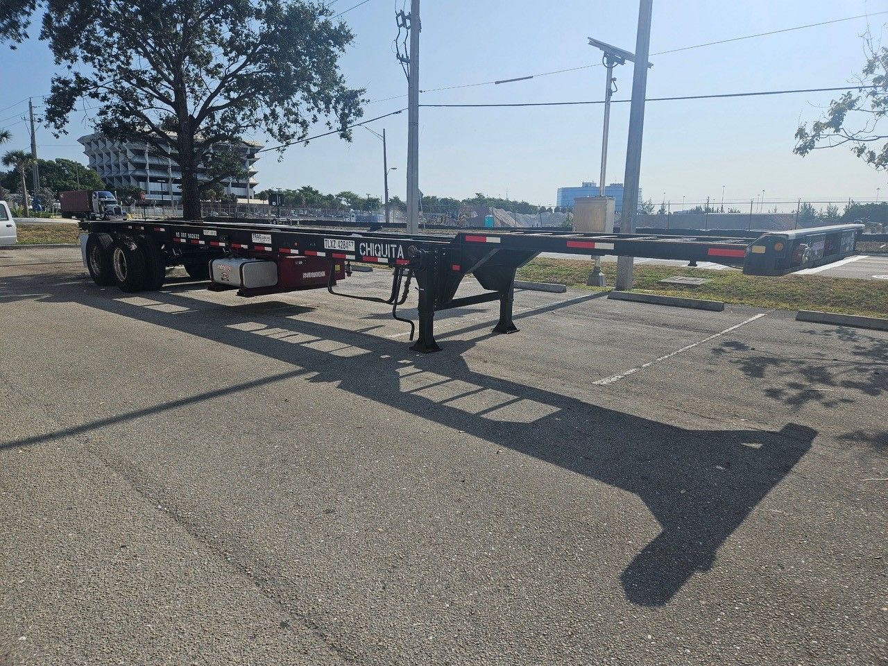 Empty black flatbed trailer parked on an asphalt lot, casting a long shadow under a blue sky.