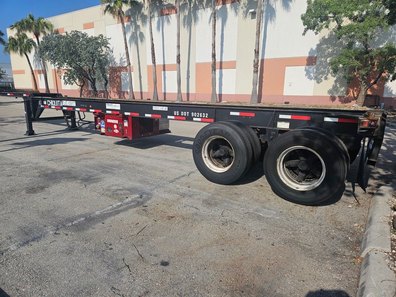 Black empty semi-trailer on a paved surface, against a building and trees.