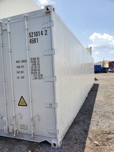 White shipping container outdoors on gravel, with details and hazard symbol visible. Blue sky in background.