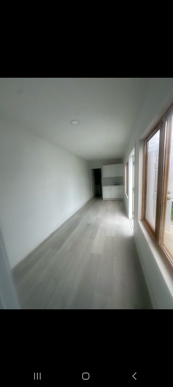 Empty interior of a long room with light-colored flooring, white walls, and large windows on one side.