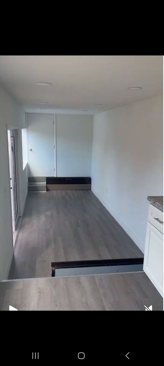 Interior of a hallway with white walls and wooden flooring, leading to a door.