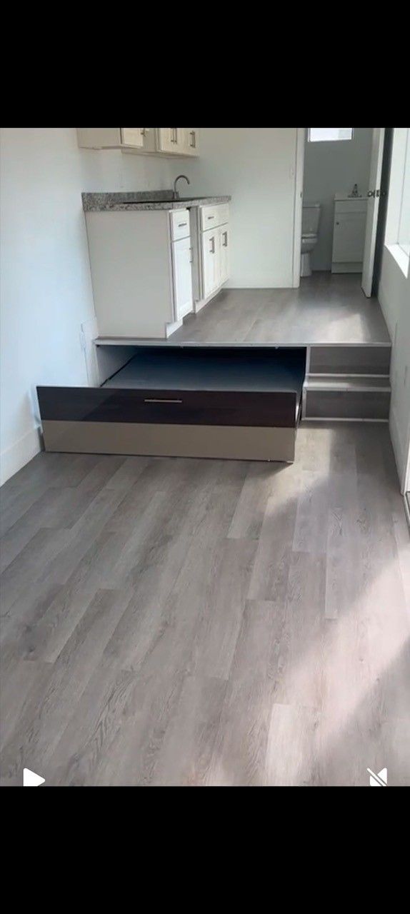 A small apartment with a raised platform for a kitchen and a pull-out bed. Gray floors, white cabinets.