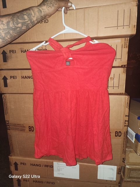 Red halter-neck top hanging on a white hanger, held by a person. Stack of boxes in background.