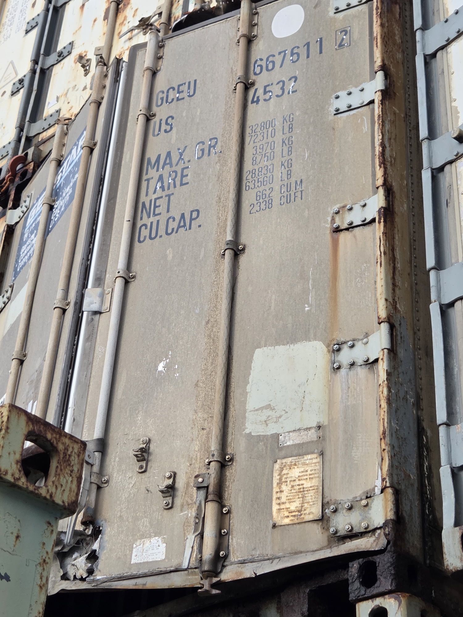 Weathered shipping container, markings visible. Gray with rust, vertical door with latches.