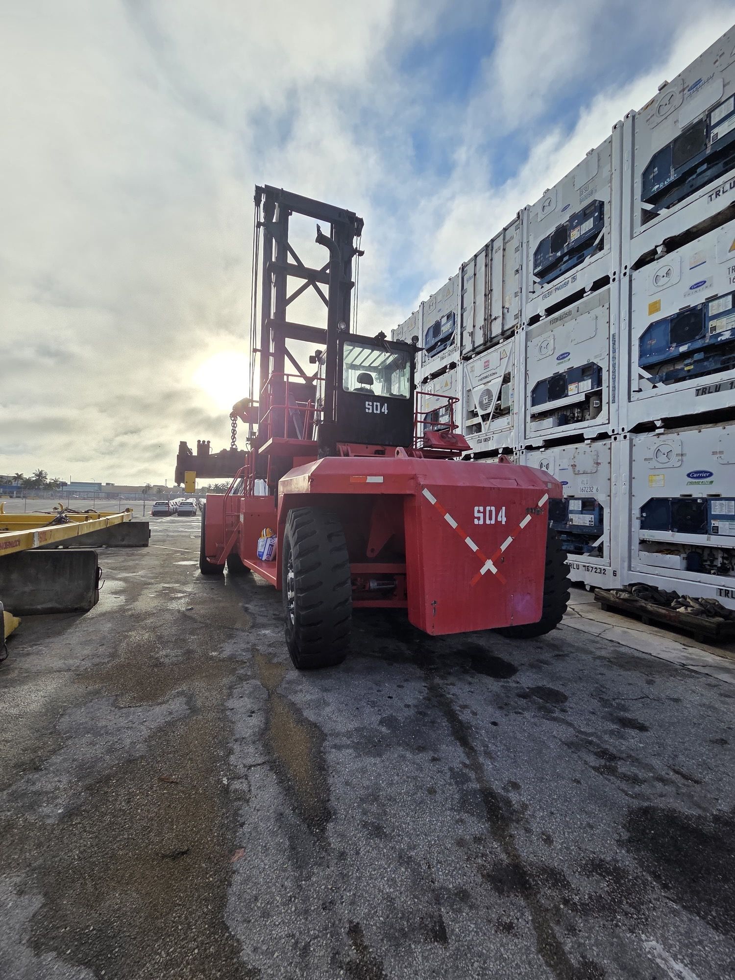 Red container handler at a port, numbered 504, parked near stacked white containers on a cloudy day.