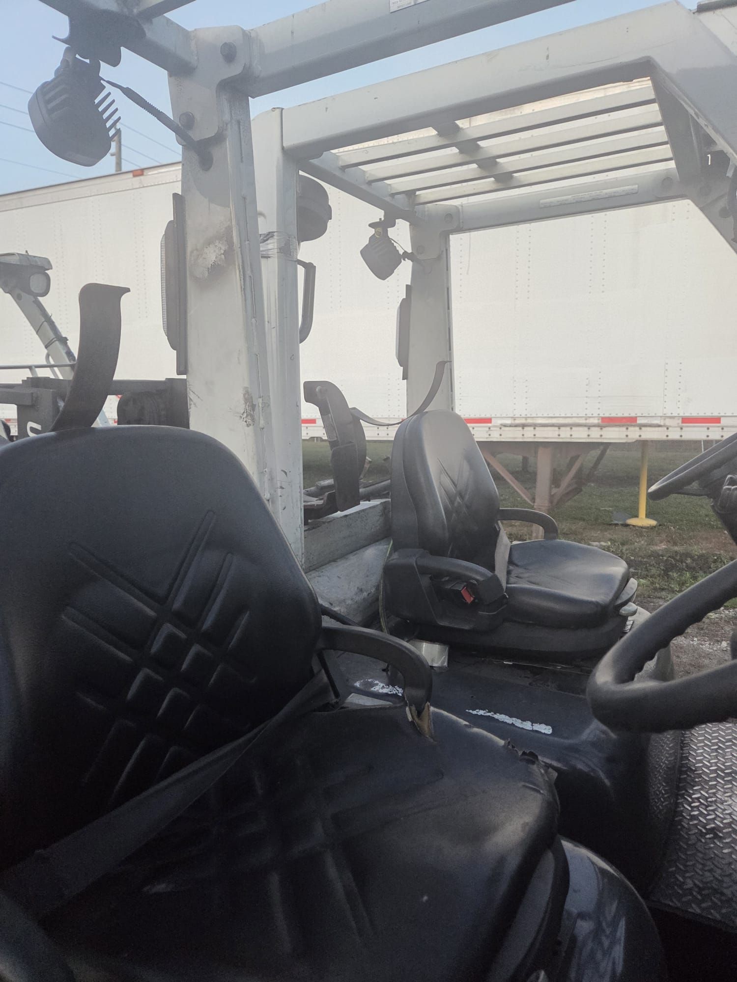Interior of a forklift cab with black seat, steering wheel, and overhead protection, parked in front of a white trailer.