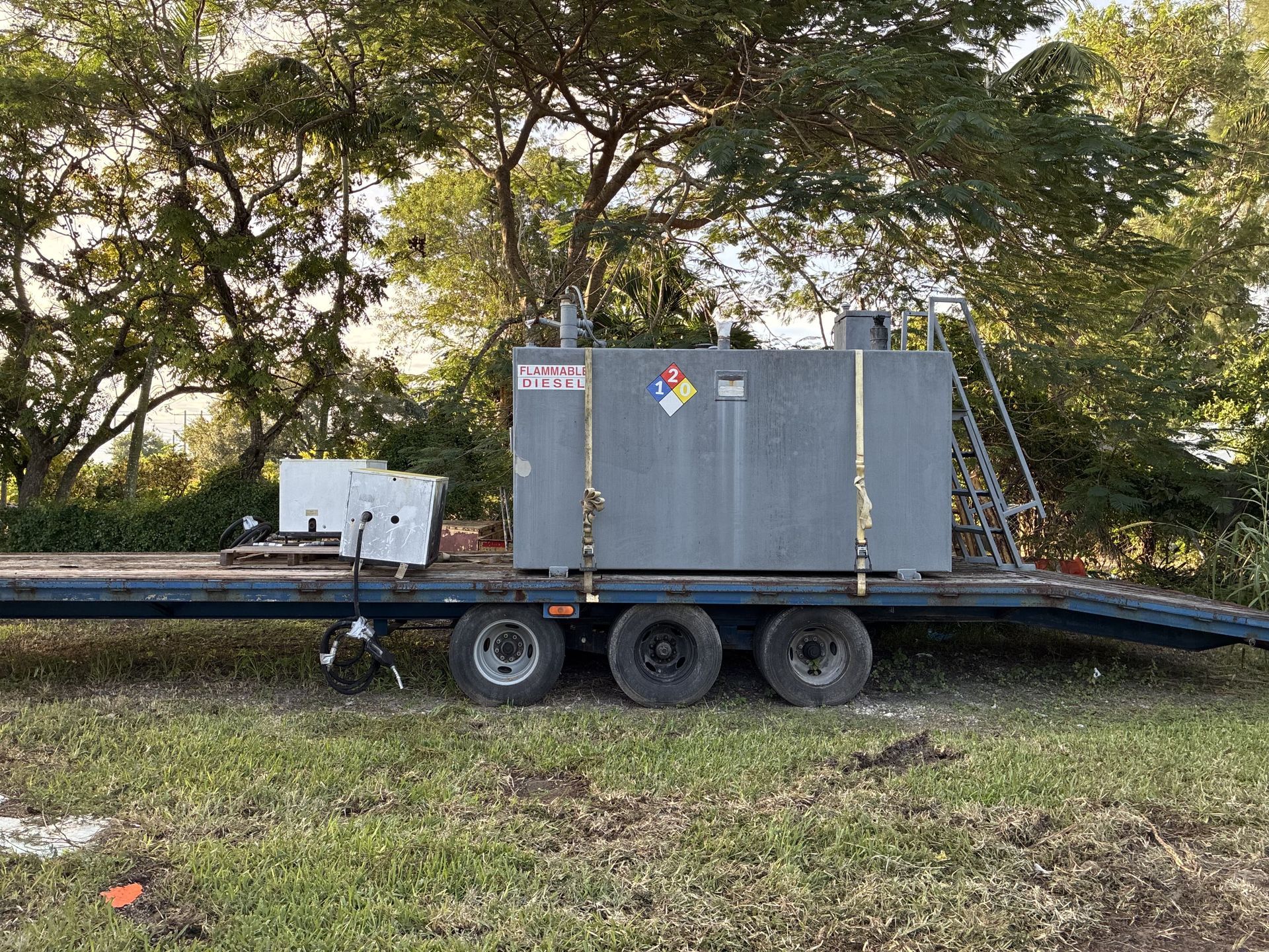 A large gray tank on a trailer, secured with straps. The tank has hazard labels and is outdoors near trees.